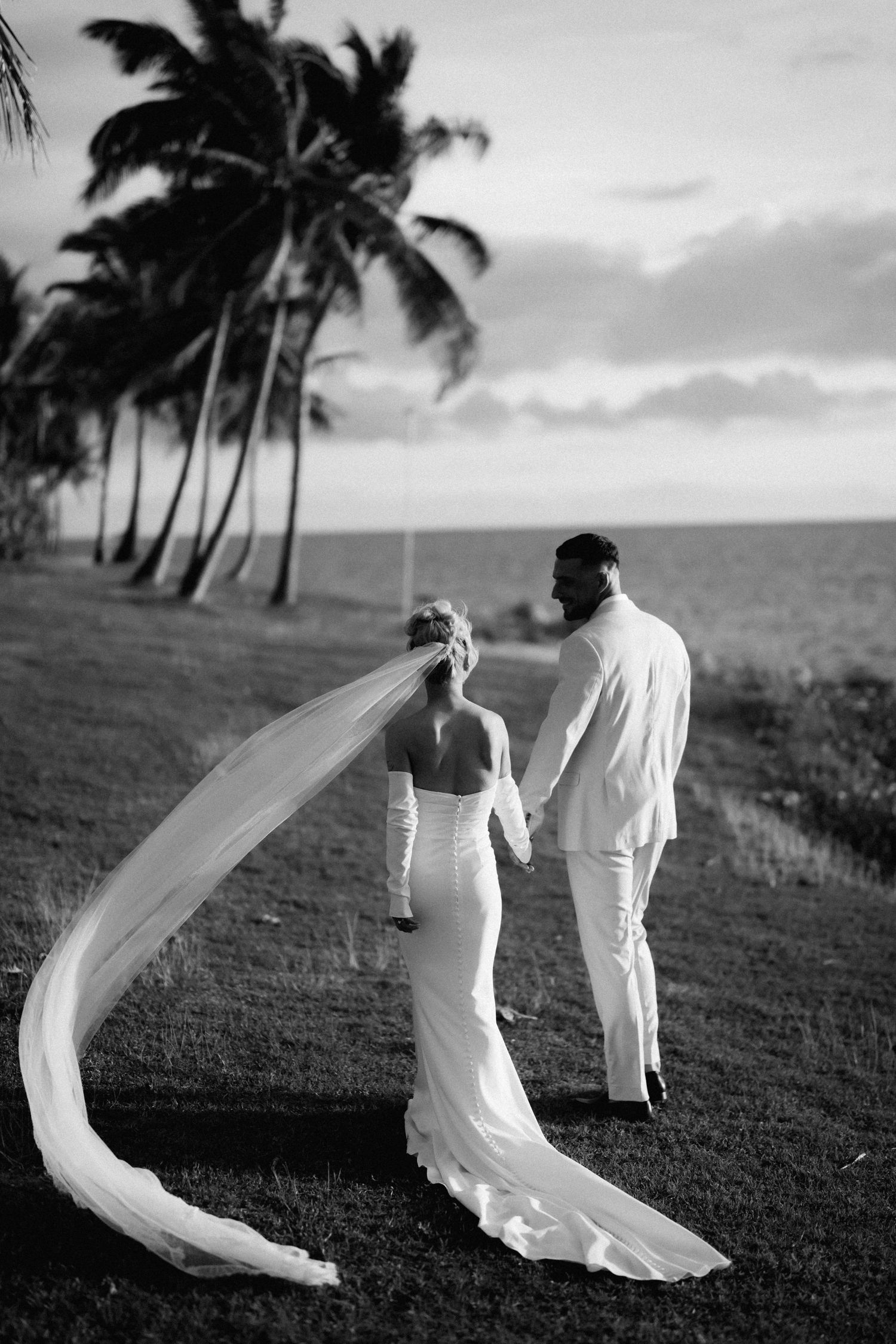 Bride and groom stand oceanside with flowing veil and palm trees in black and white.