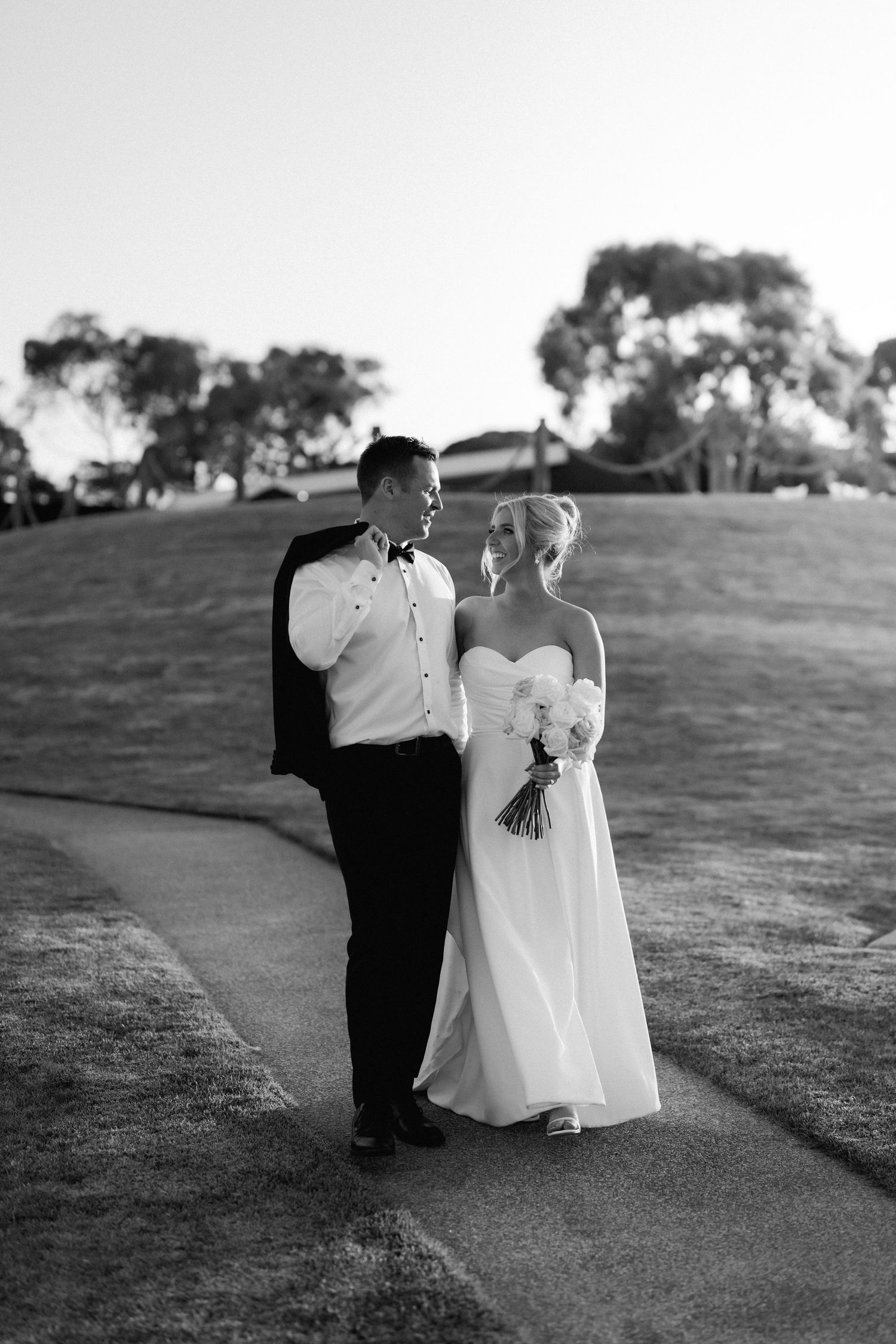 A romantic black and white portrait of newlyweds standing on a path with trees in background.