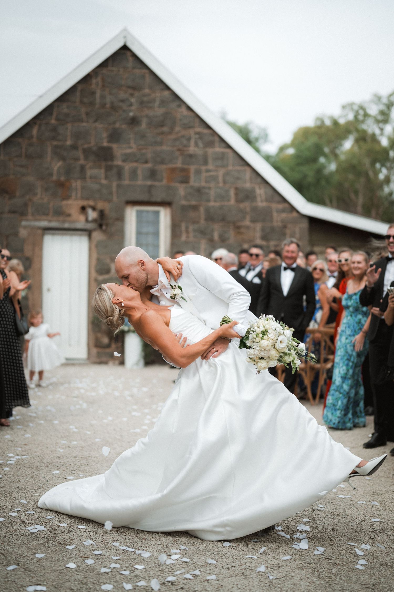 Romantic wedding dip kiss in front of a rustic stone church with guests watching and confetti falling.