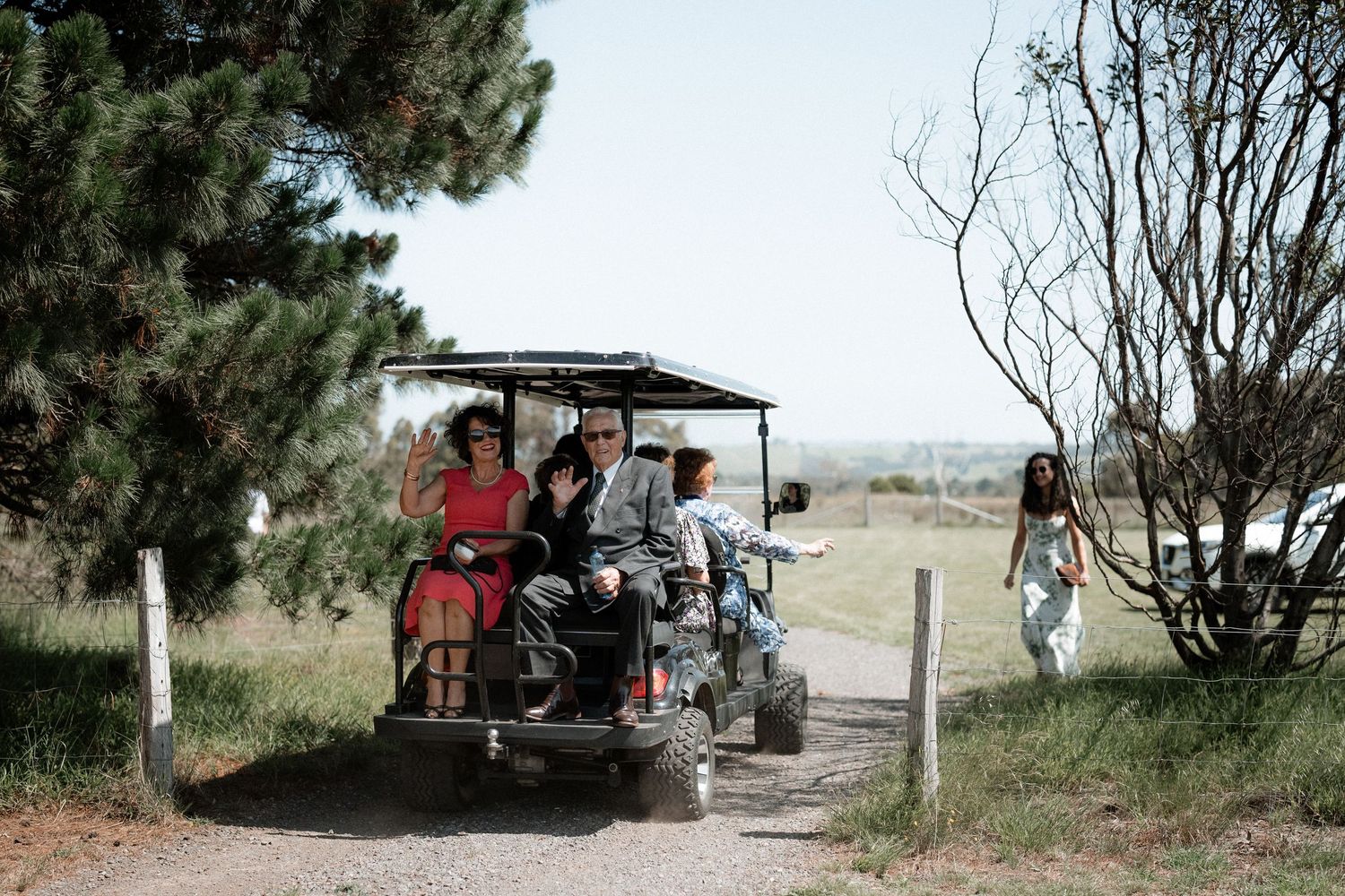 A group of people ride in a golf cart along a dirt path at a rural outdoor location on a sunny day.