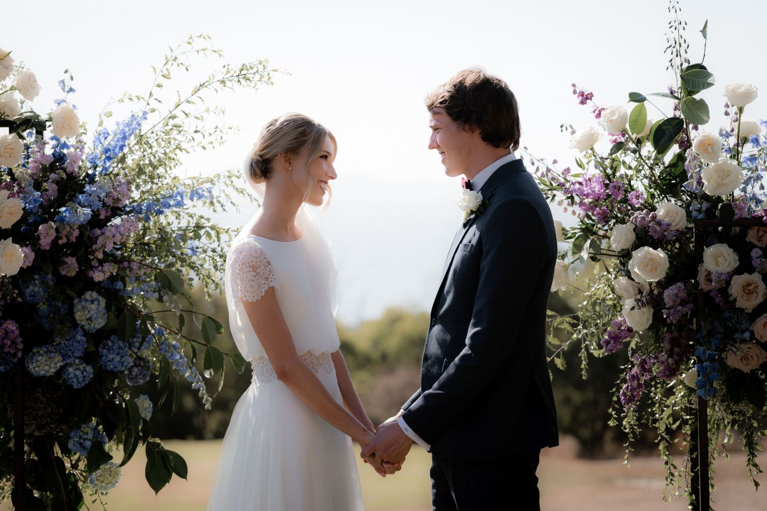 A couple holds hands at wedding ceremony with floral arch beautifully decorated in blue and purple flowers during sunset.