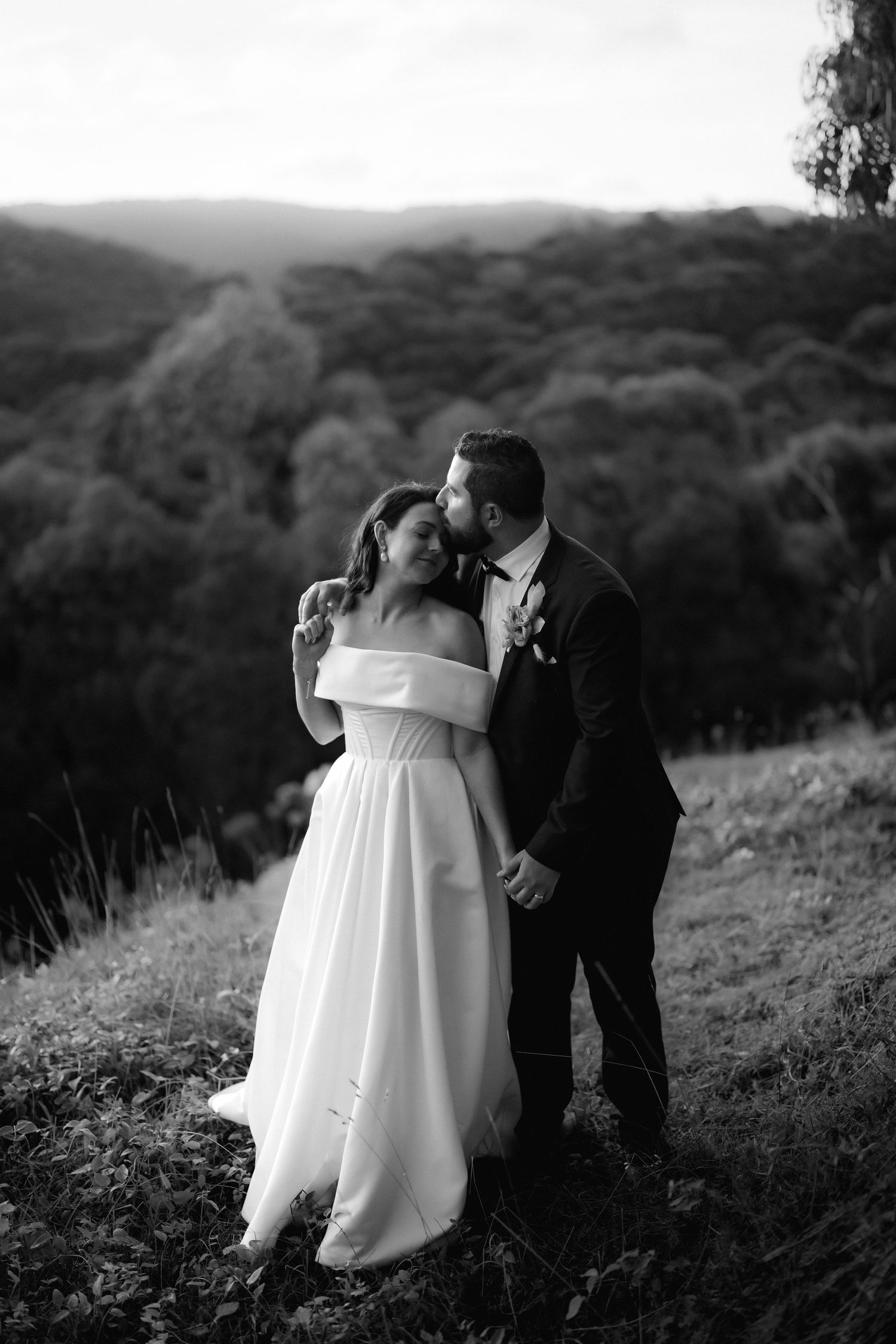 Black and white wedding photo of couple embracing on hillside with scenic mountain landscape in background.