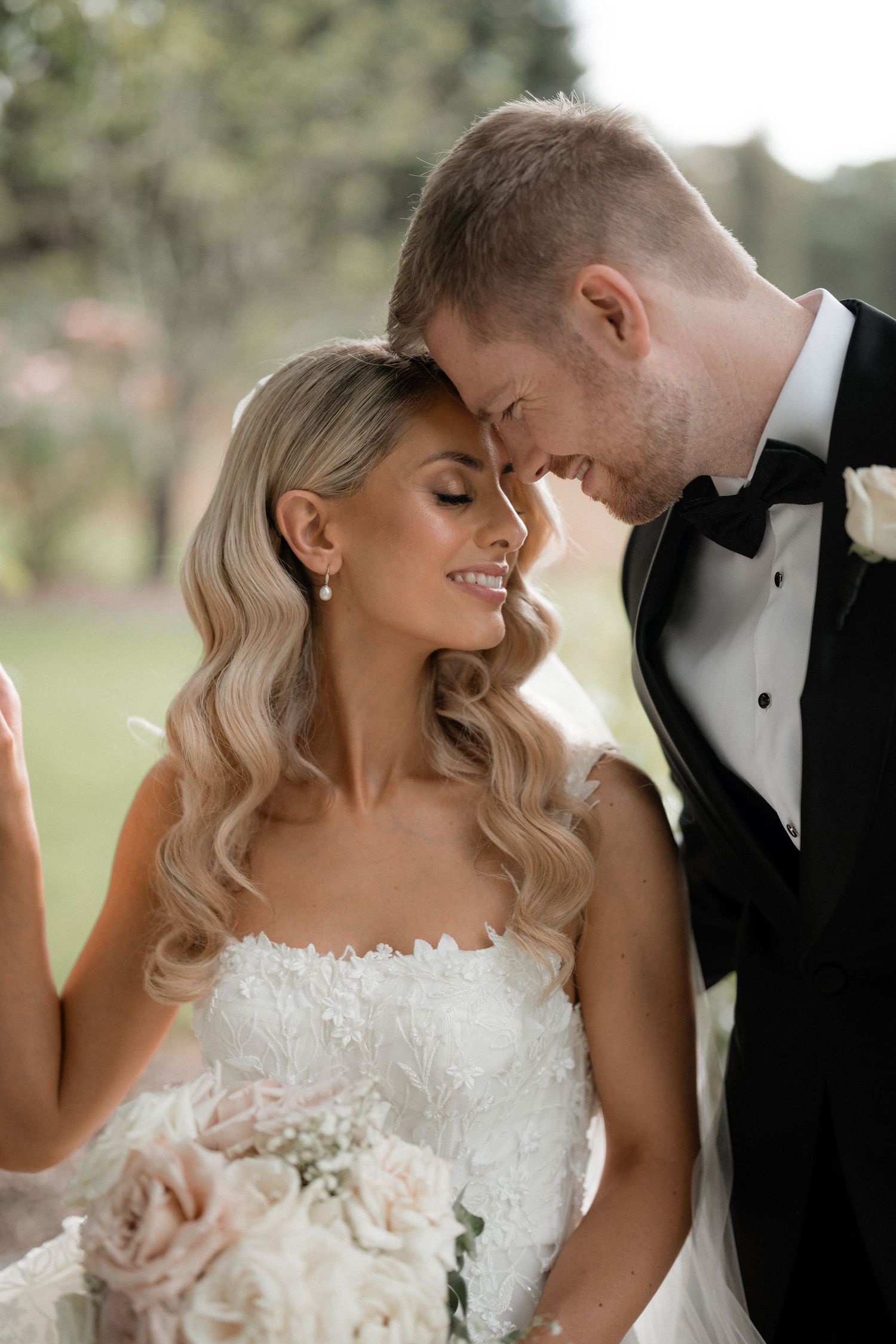 A romantic wedding photo of a bride in a white lace gown and groom in tuxedo sharing an intimate moment outdoors.