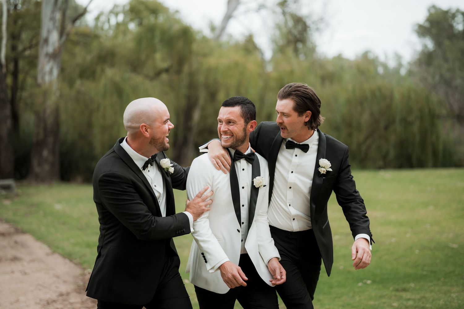 Groomsmen in black tuxedos and bowties walk together outdoors while laughing and smiling during wedding celebration.