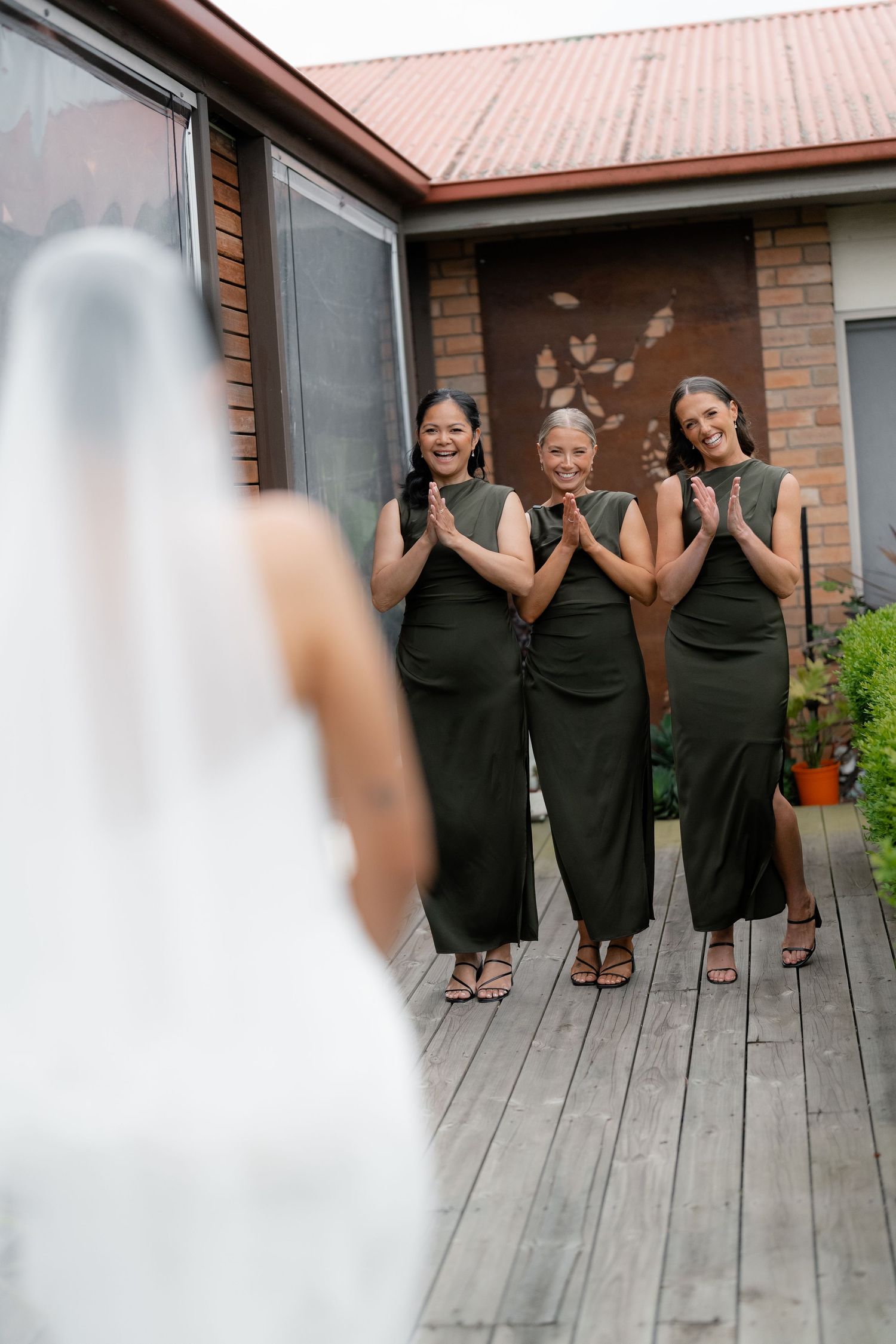 Three bridesmaids in matching black dresses pose playfully on a wooden deck outside a brick building.