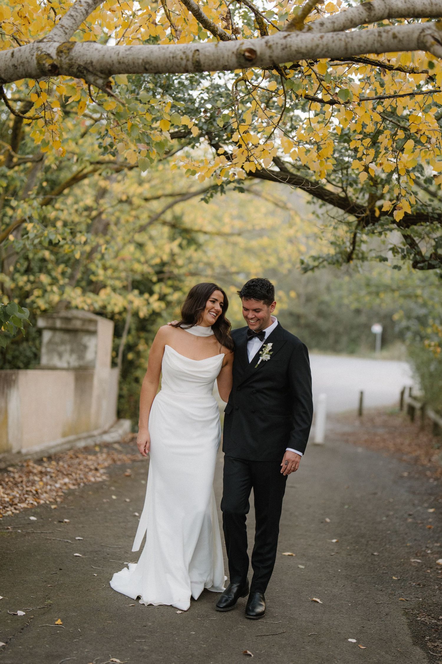 A couple in wedding attire walking through a path with autumn trees and yellow leaves overhead.