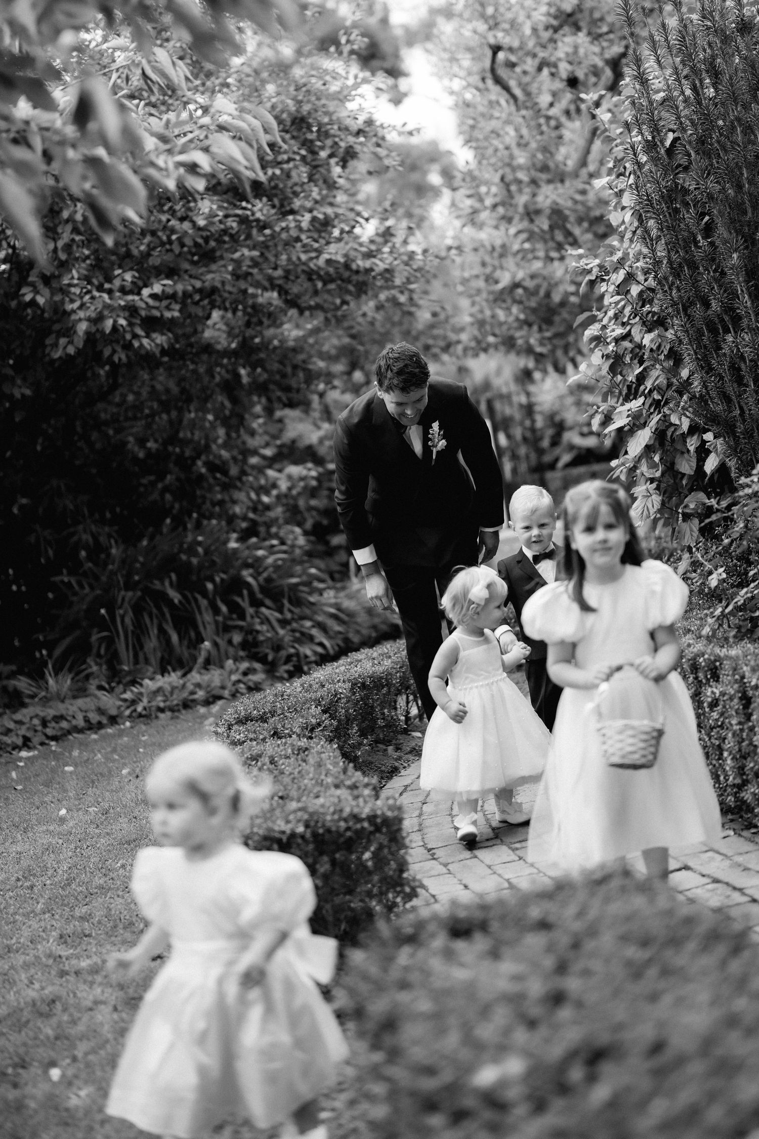 Black and white photo of children in formal attire playing in a garden setting during a wedding.
