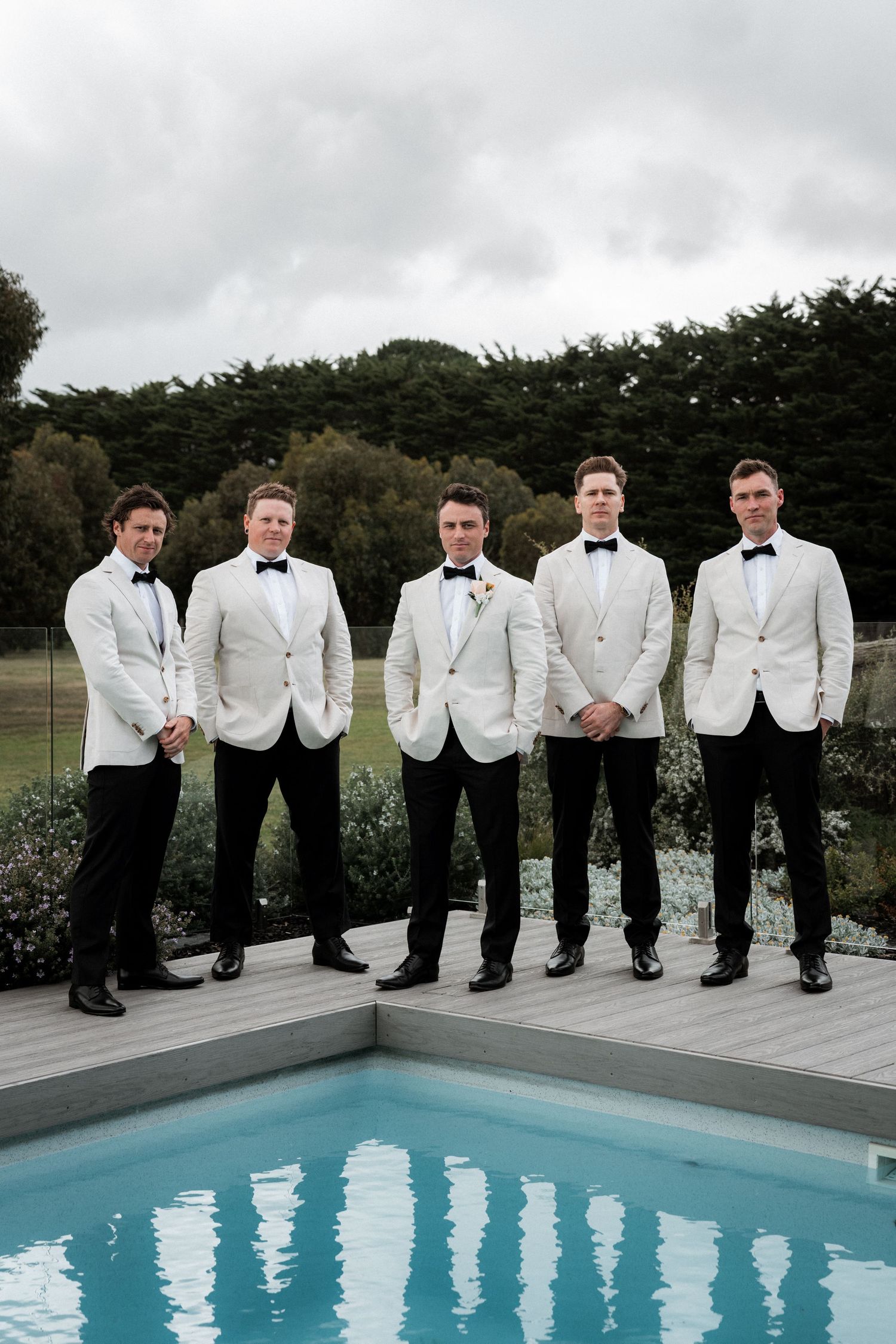 Groomsmen in white dinner jackets and black pants pose together by a swimming pool on an overcast day.