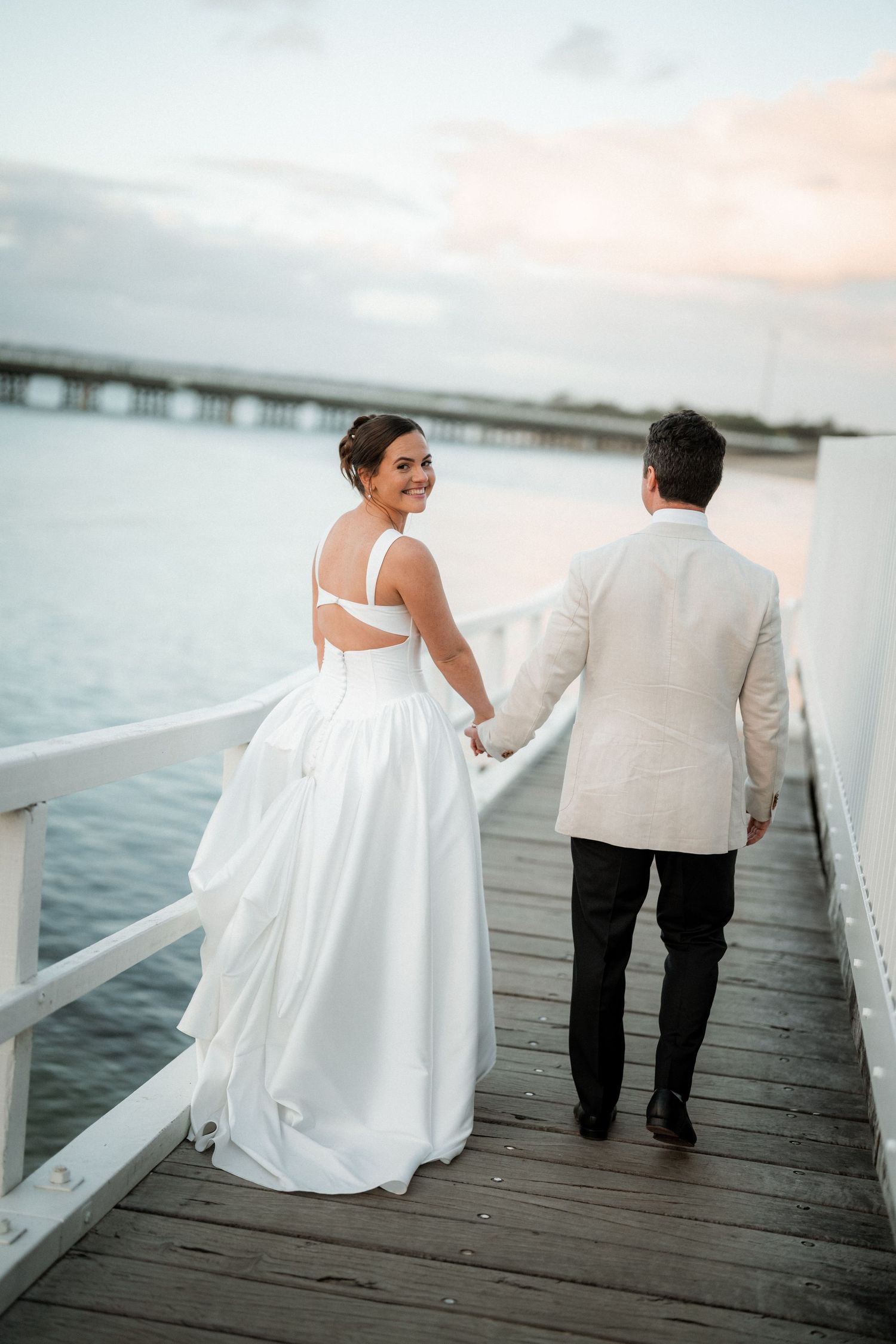 A couple in wedding attire walks along a waterfront dock during sunset.