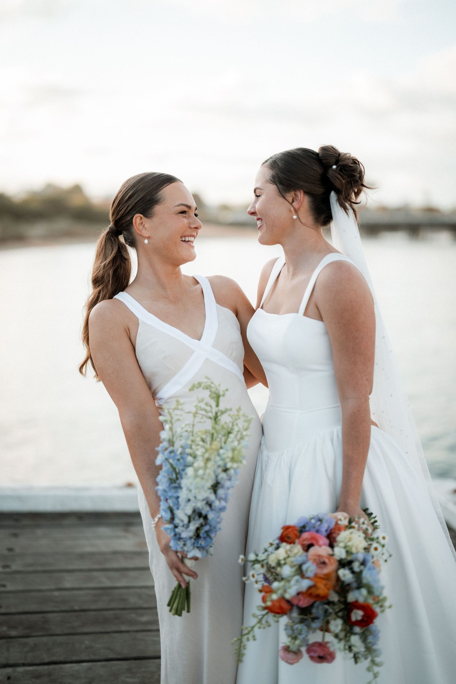 Two brides share a joyful moment on a dock at sunset, holding beautiful bouquets of blue and coral flowers.
