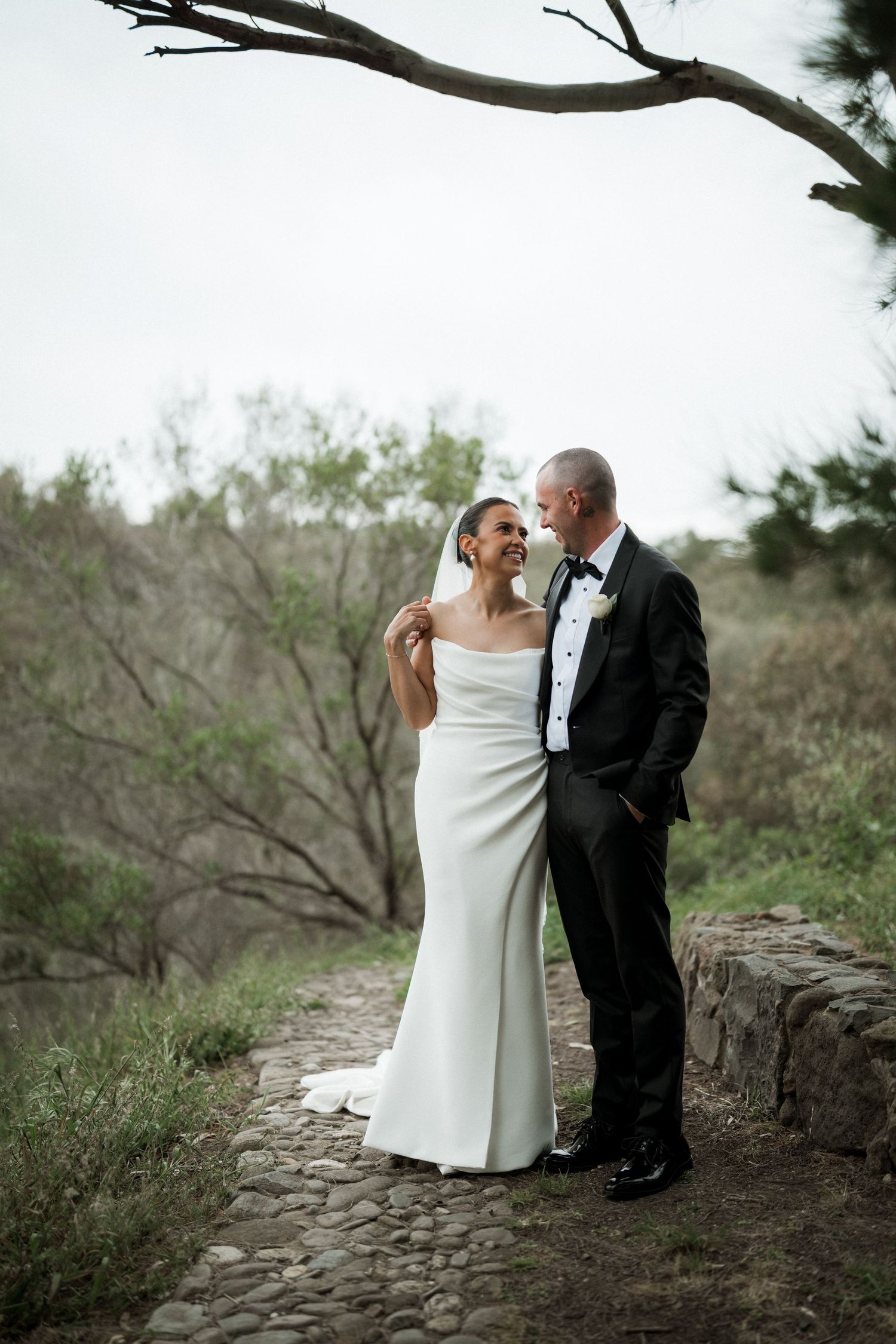 A romantic wedding portrait of a couple standing on a stone path under a dramatic tree branch.