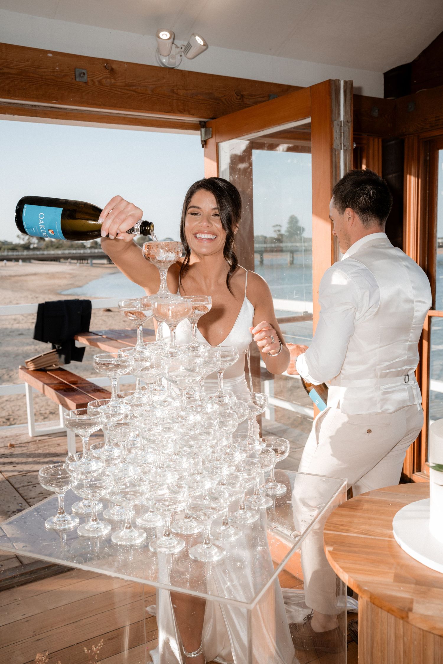 People in white attire pour champagne into a tower of crystal glasses.