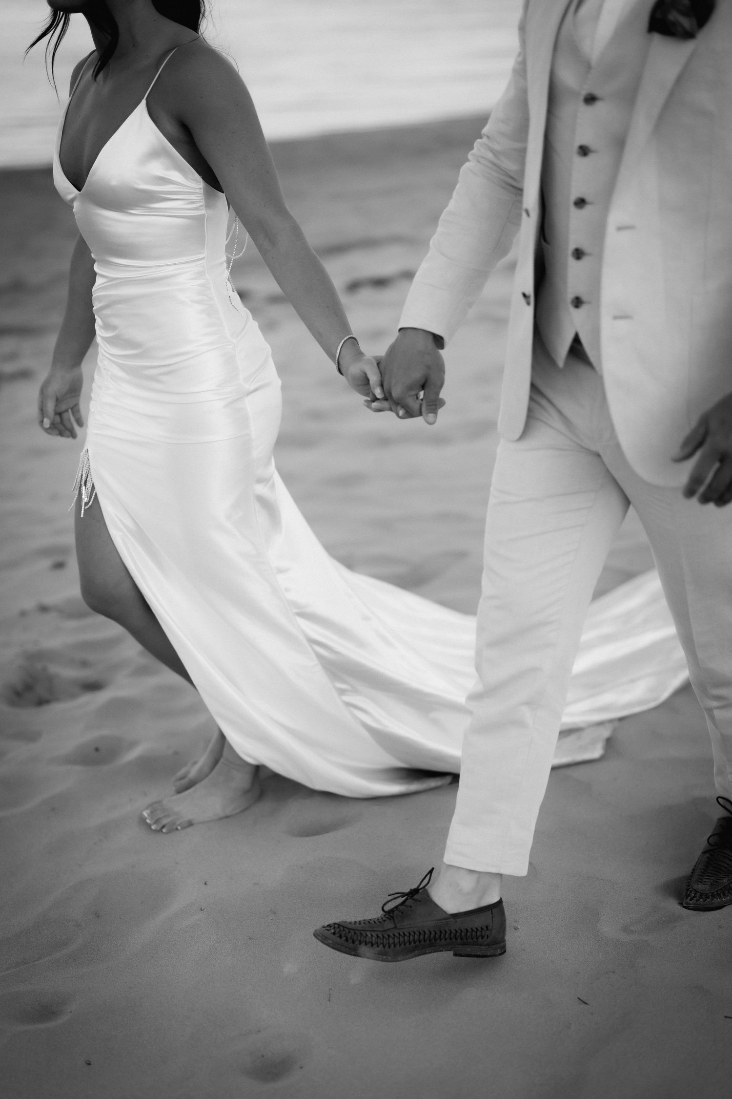 Black and white photo of newlyweds holding hands while walking barefoot on beach sand.
