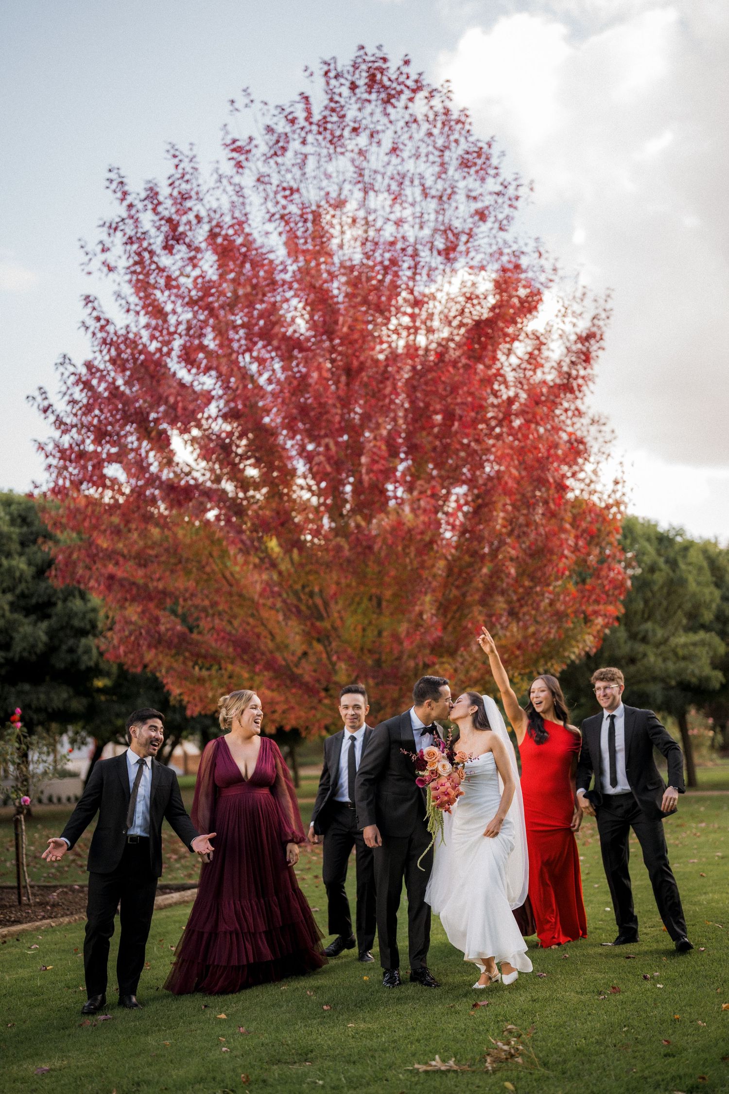Wedding party walks outdoors near a stunning red autumn tree, bride and groom kissing while guests celebrate around them.