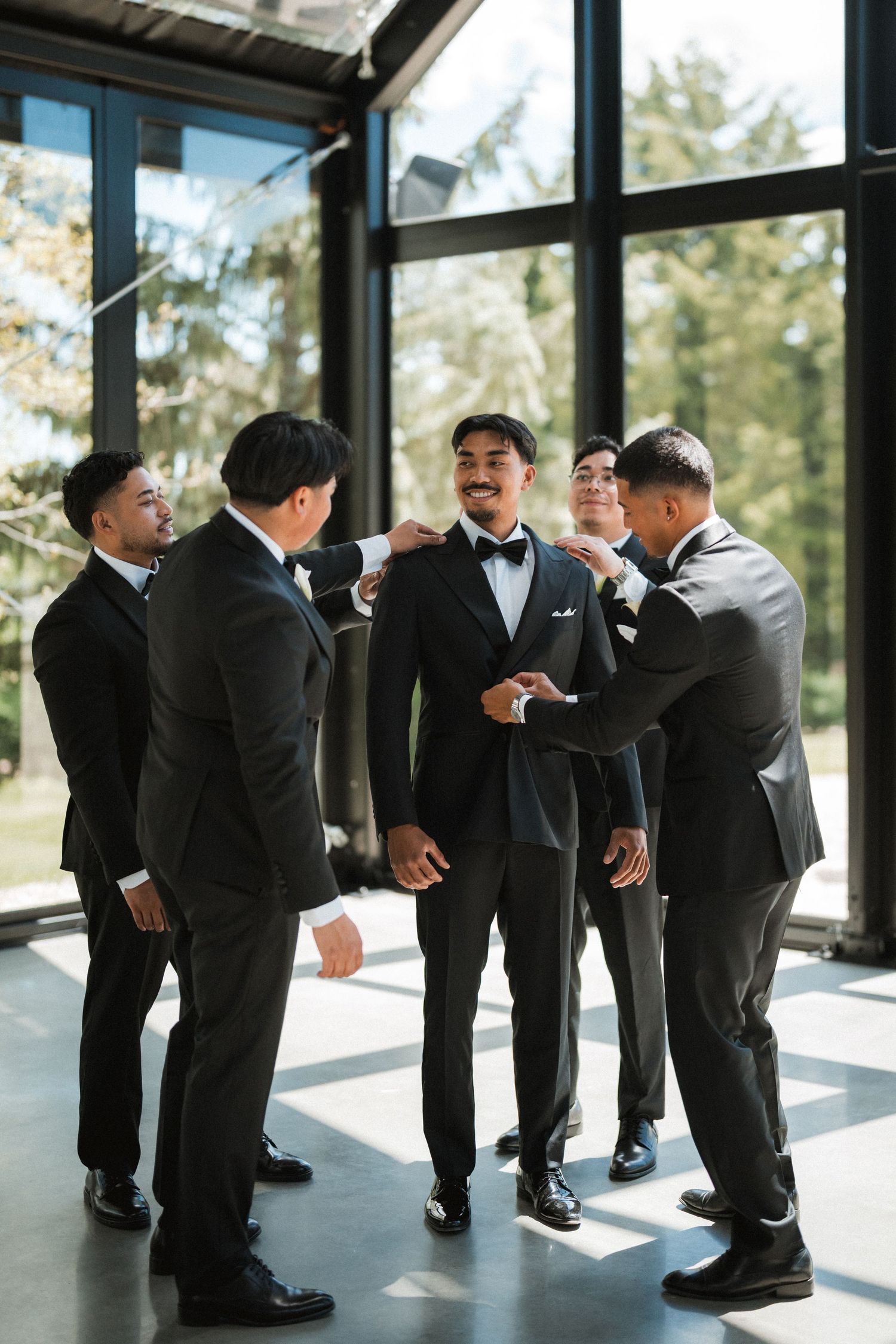 Groomsmen in black suits adjust the smiling groom's bow tie and jacket in a bright, sunlit room with large windows before the wedding.