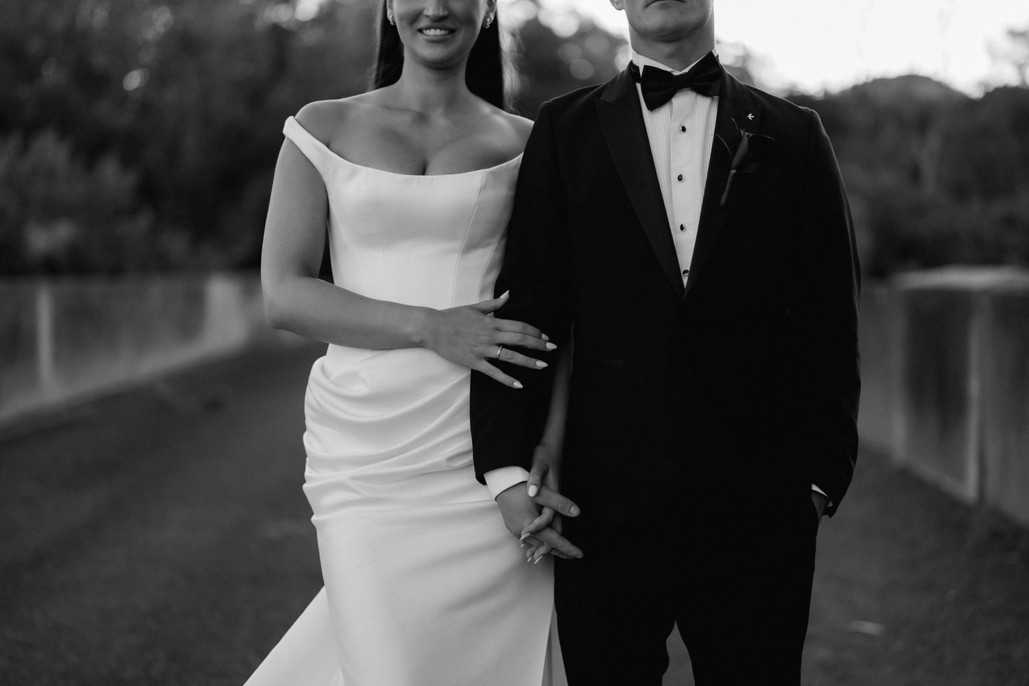 Black and white photo of a bride in an off-the-shoulder gown and a groom in a tuxedo and bow tie holding hands outdoors.