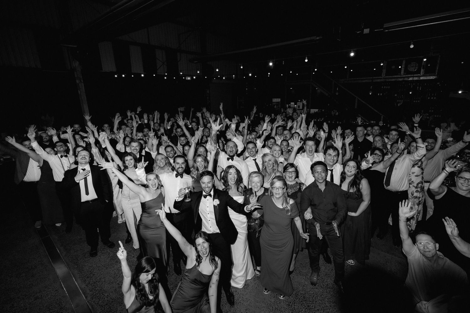 Large crowd of wedding guests dancing with hands raised during nighttime reception celebration.