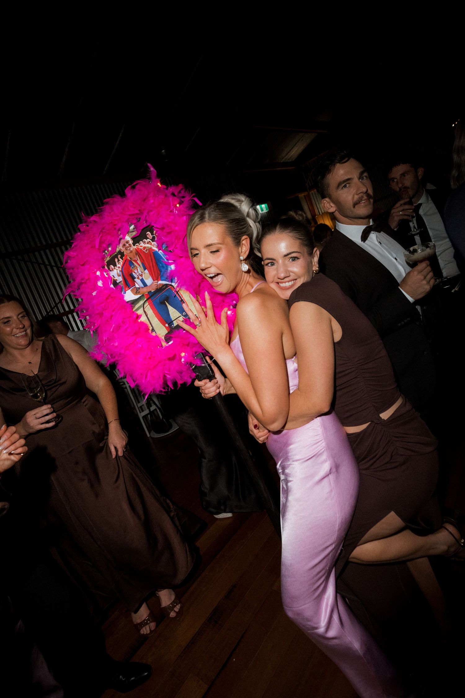 Party guests dancing and celebrating with bright pink feather boas in a dimly lit reception venue.