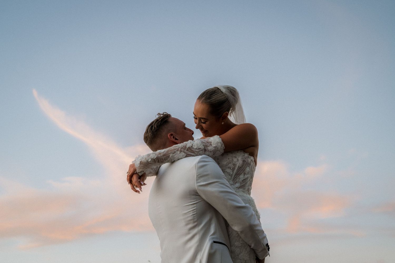 A romantic couple embraces against a pastel sunset sky during their wedding celebration.