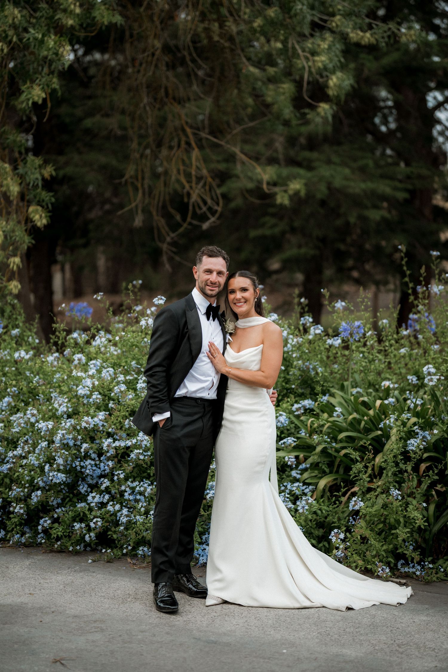 Wedding couple poses together on garden path surrounded by blooming blue flowers and greenery.