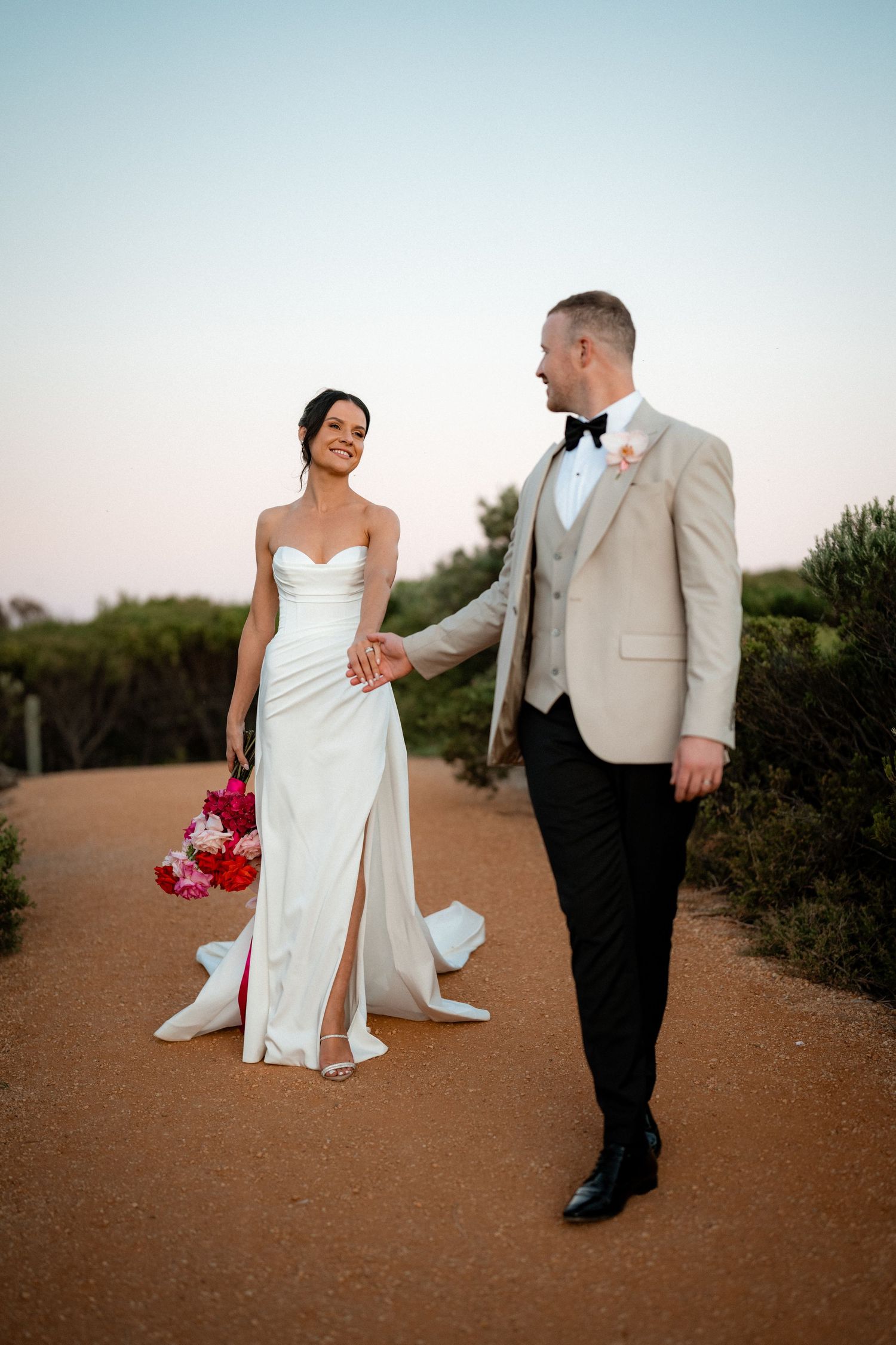 A wedding couple poses playfully on a desert path with pink bouquet at sunset.
