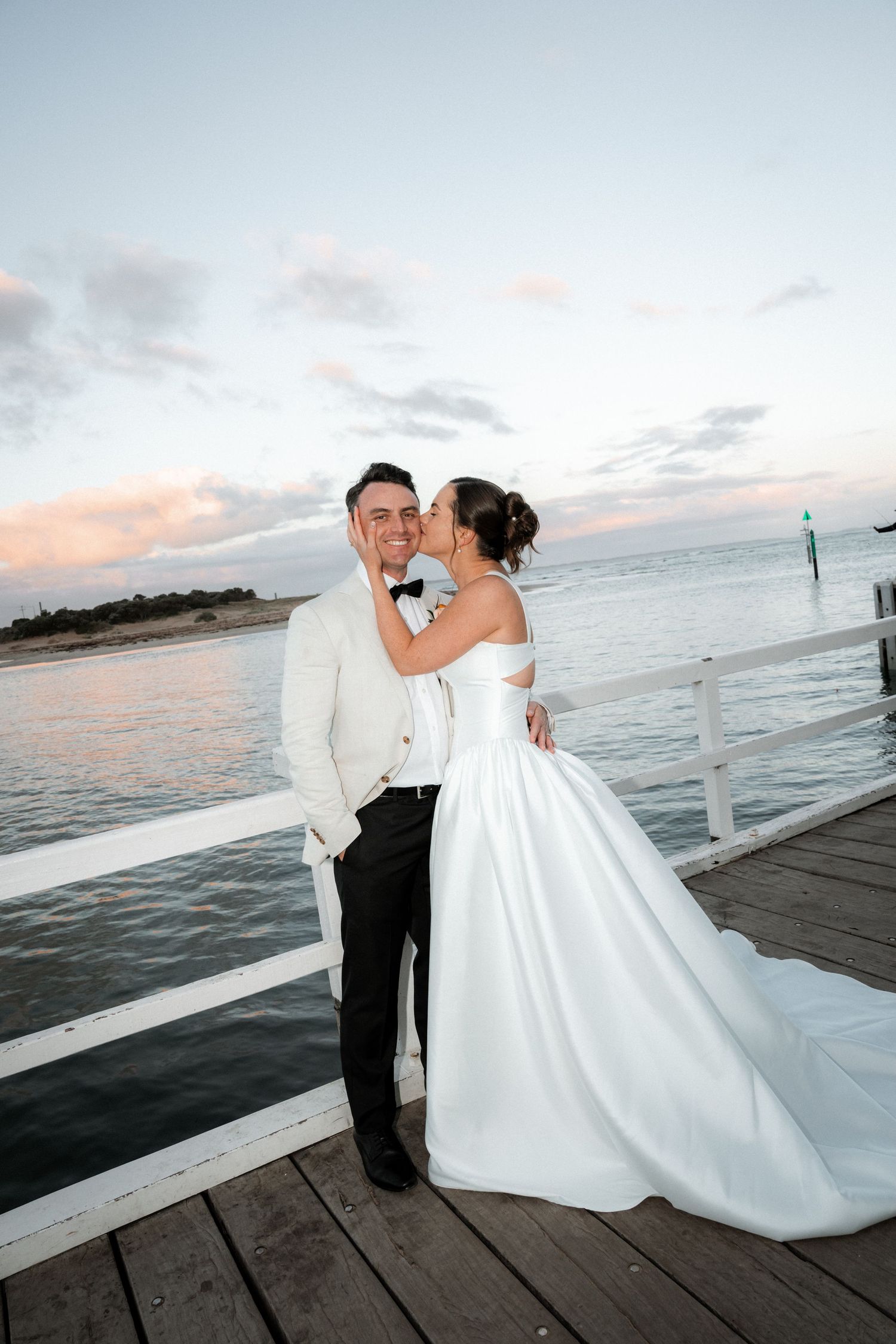 A couple in wedding attire embraces on a wooden pier at sunset with ocean views in the background.