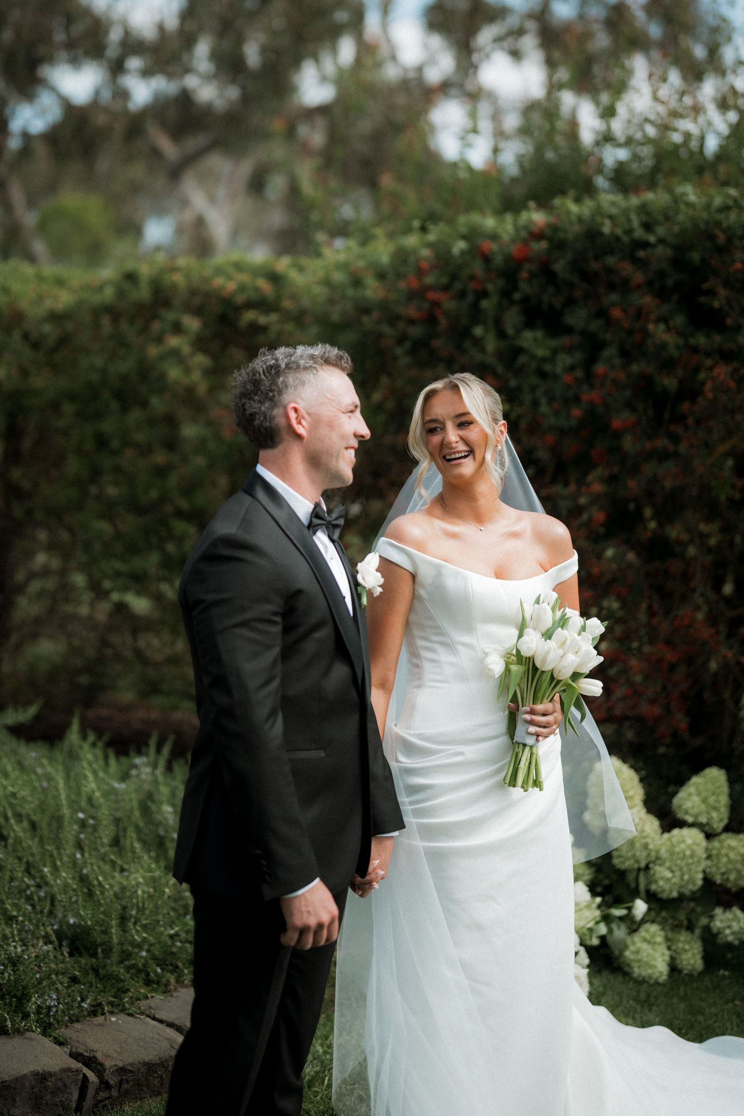 Wedding couple shares joyful moment in garden setting with white flowers and greenery.
