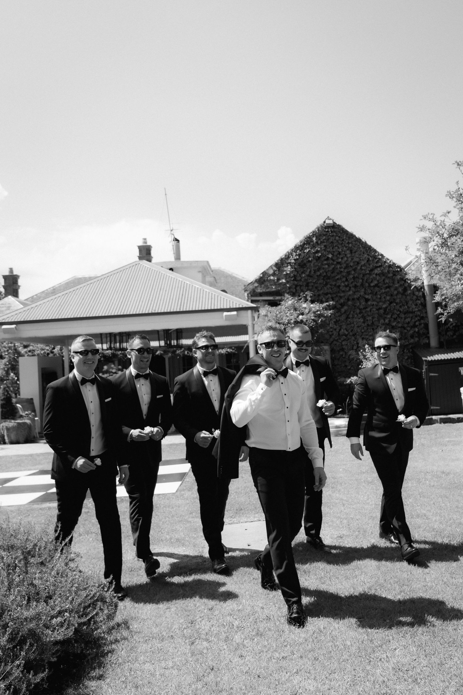Groomsmen in black tuxedos walk together on grass in a casual formation during wedding photos.