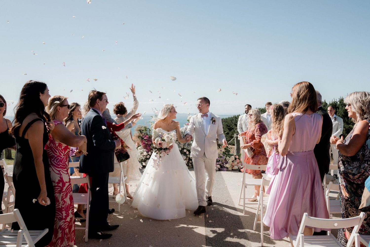 Wedding guests in formal attire celebrate during an outdoor ceremony against a blue sky.
