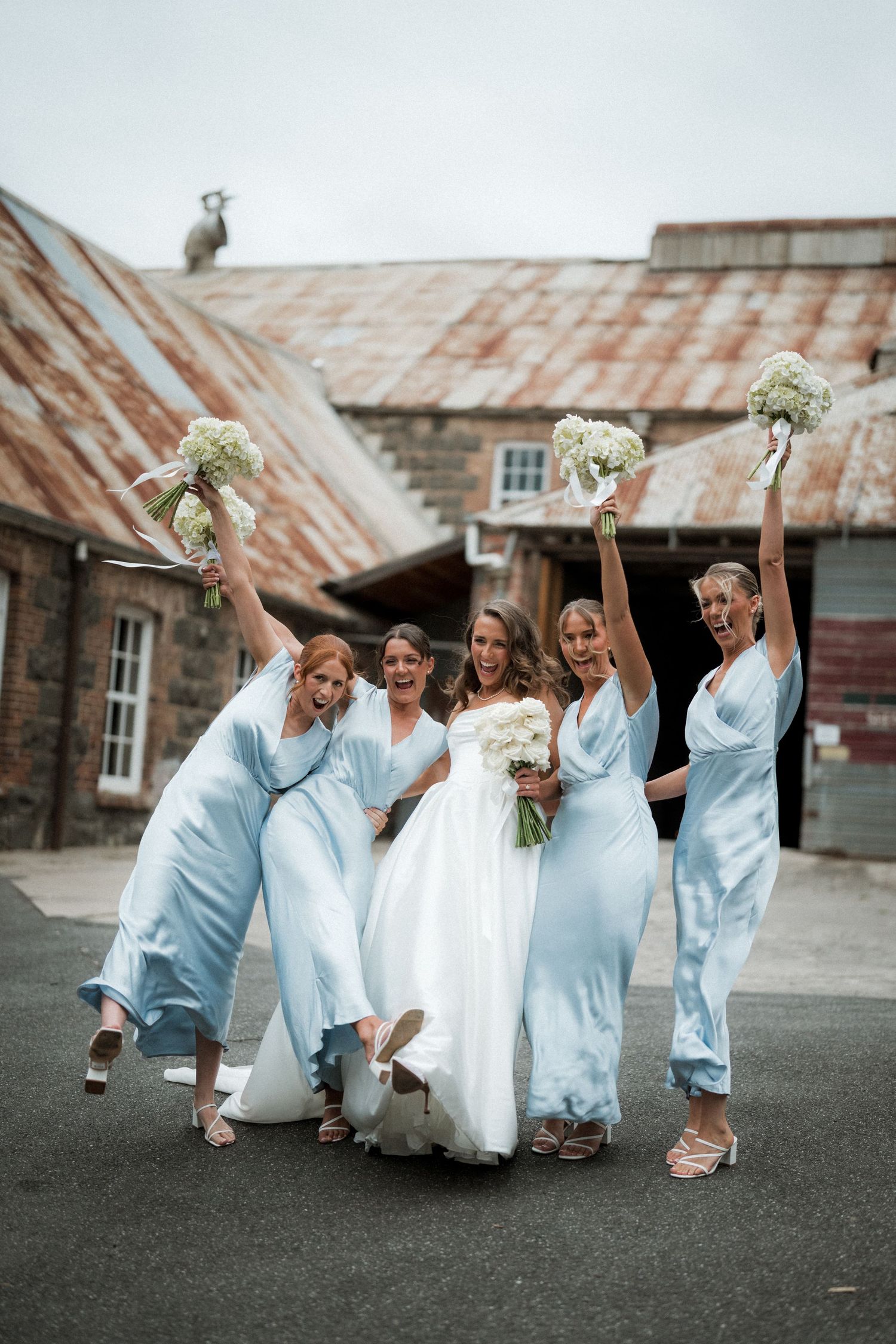 Bridesmaids in light blue dresses strike playful pose with bride in white gown against rustic building backdrop.