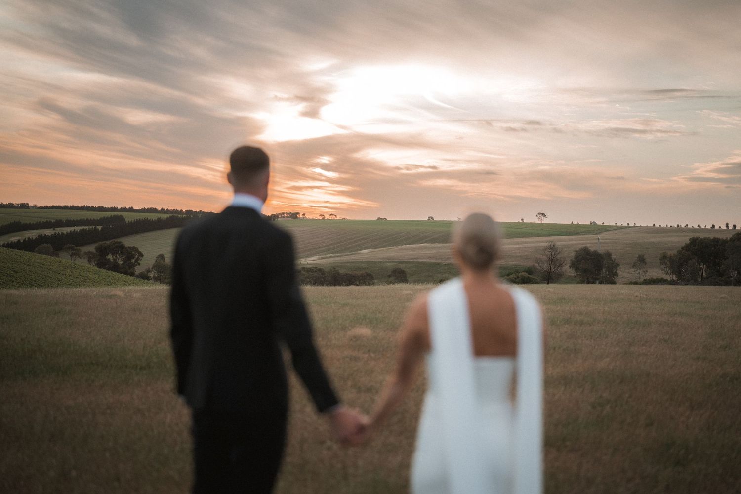 Silhouettes of a wedding couple walking hand in hand through a golden field at sunset.