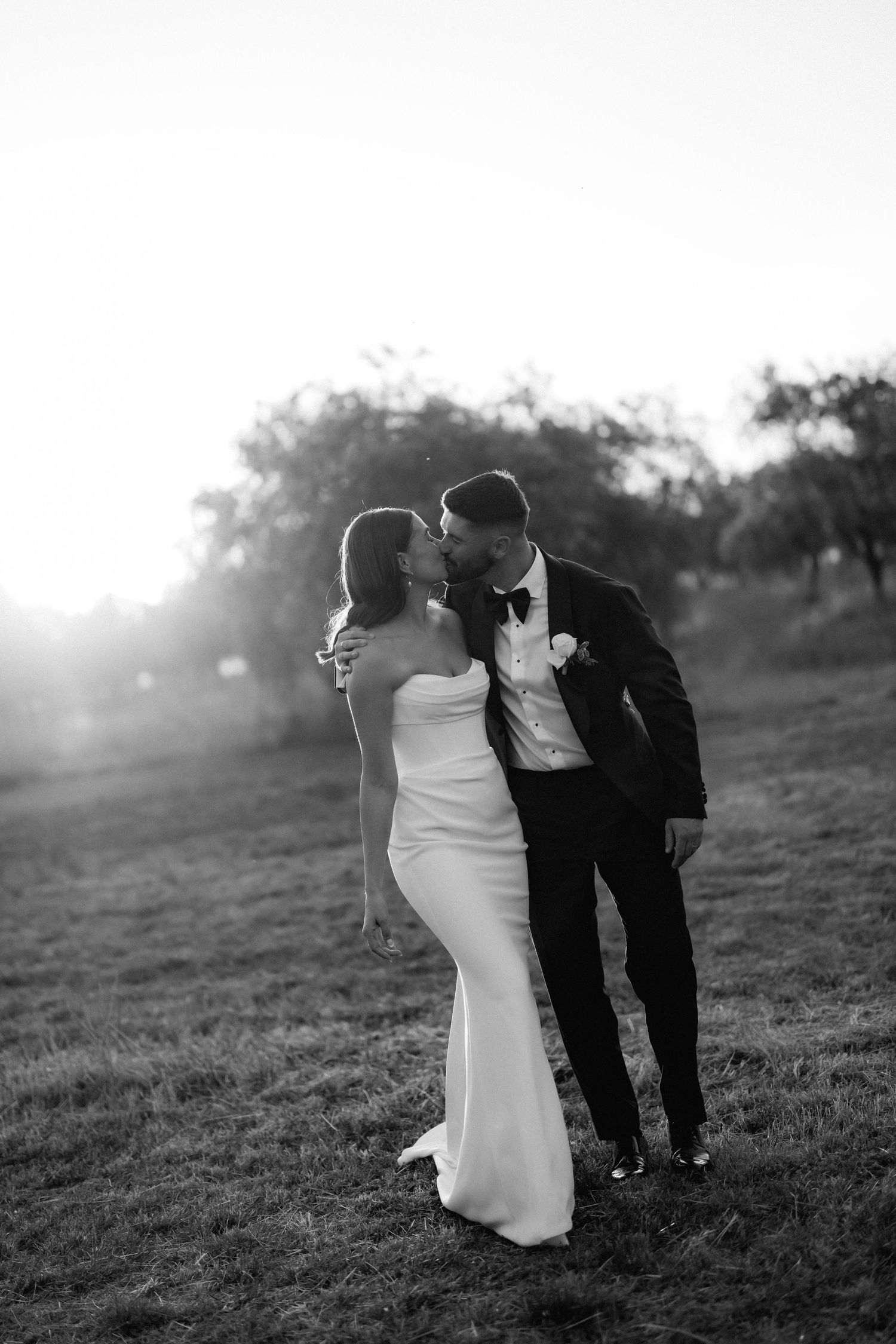 A romantic black and white wedding portrait of a couple in formal attire among rolling hills at sunset.