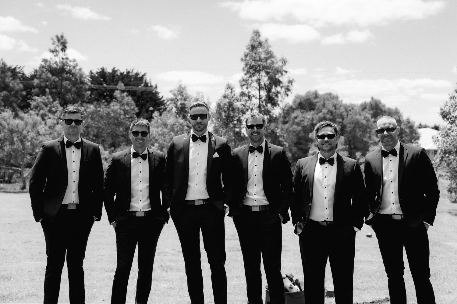 A black and white panoramic collage of groomsmen in formal suits posing outdoors against a cloudy sky.