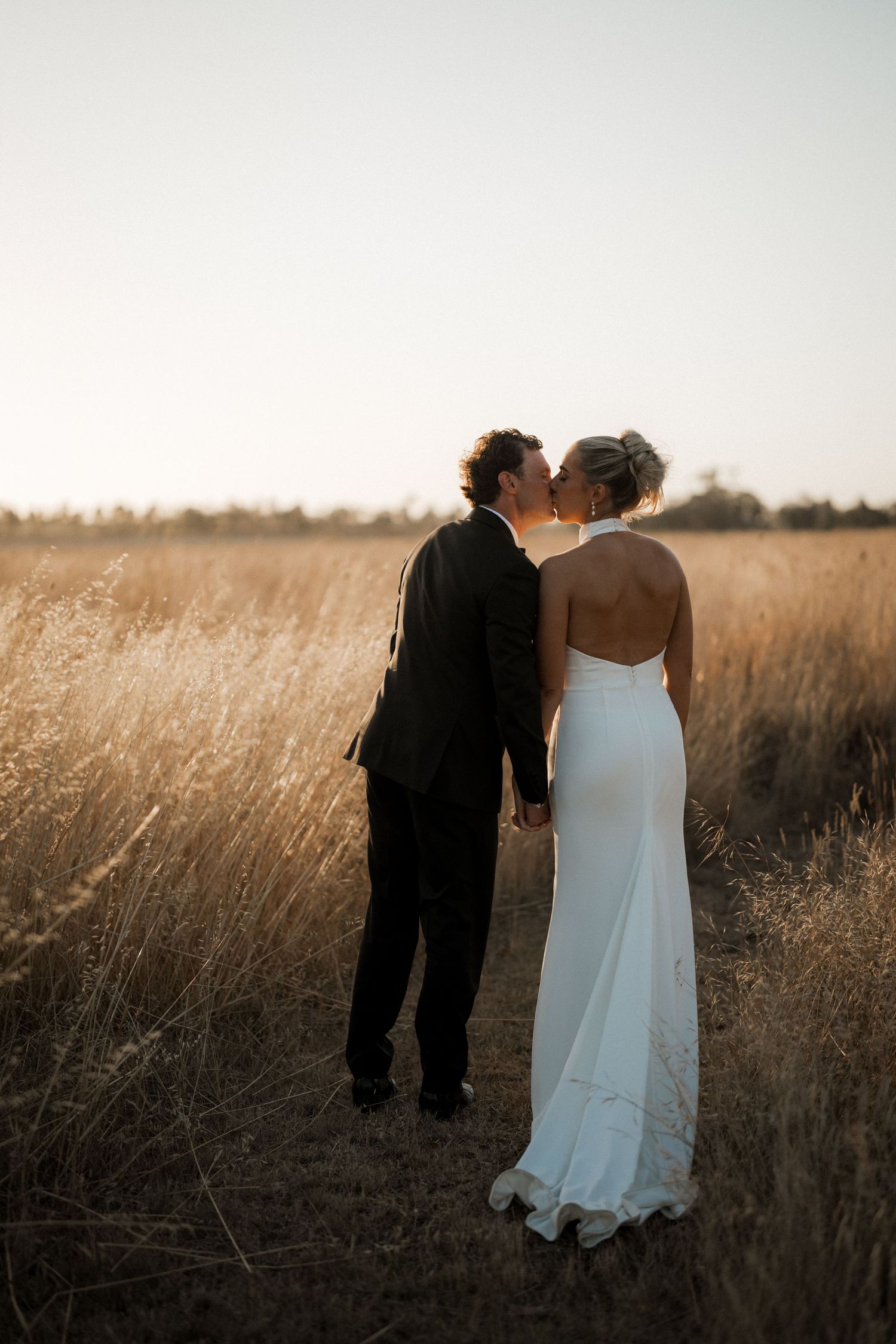 Romantic couple in wedding attire standing together in a golden wheat field at sunset.