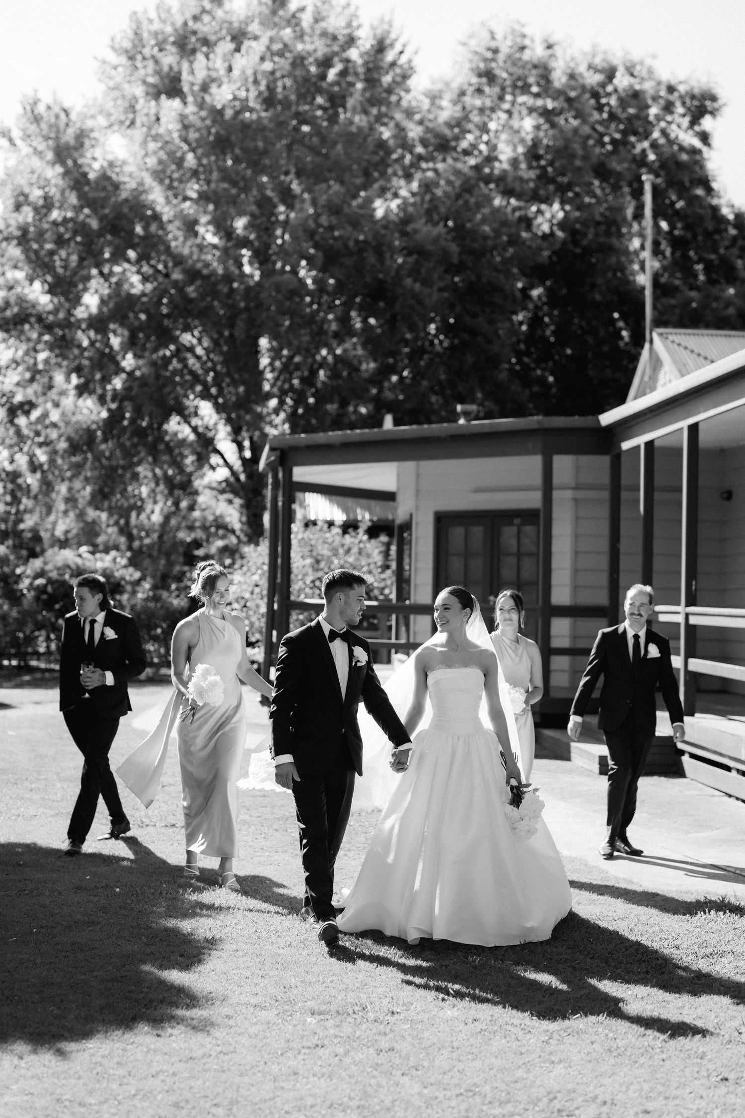 Wedding party walking together on grass path between buildings in black and white.
