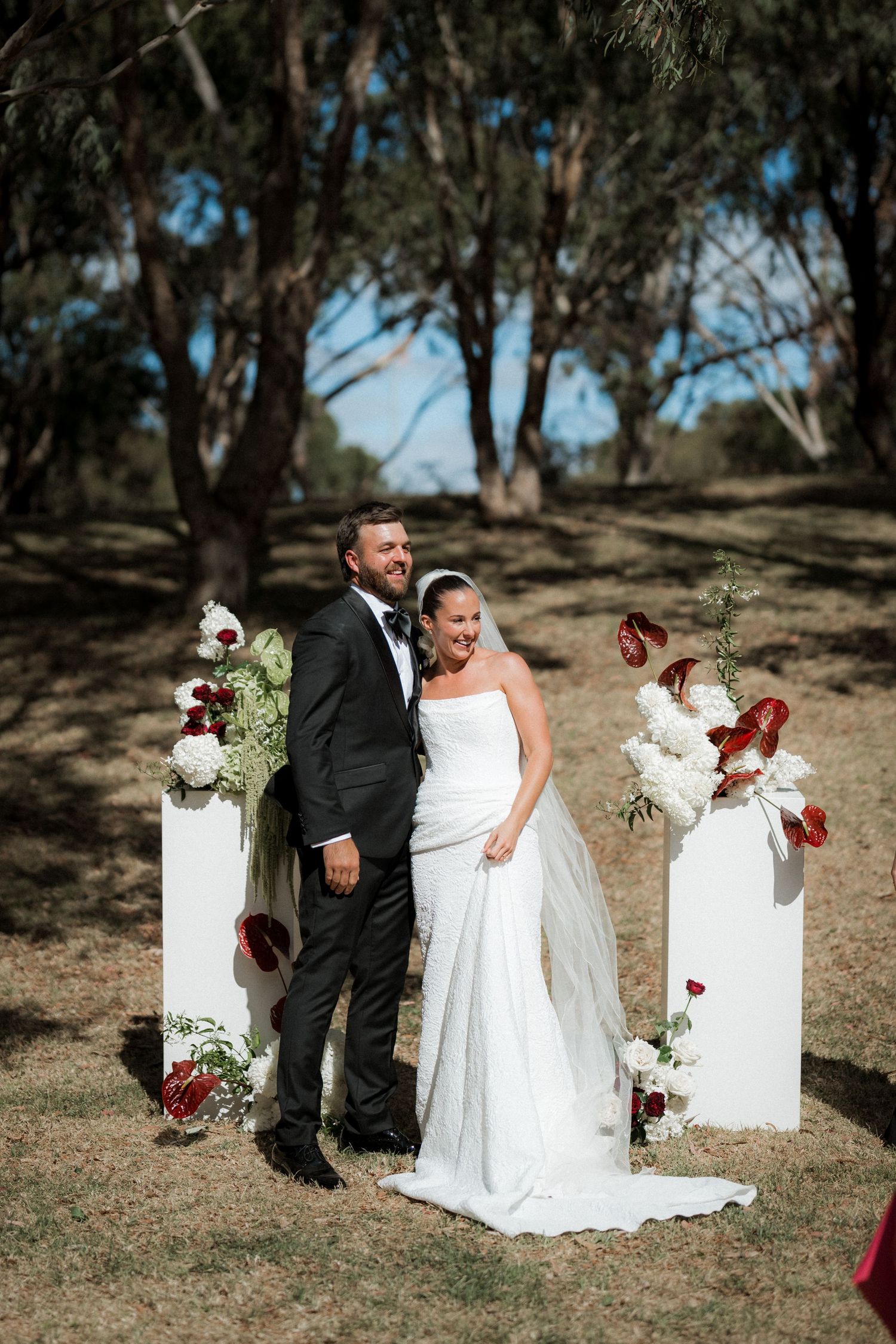 Bride and groom smiling together at outdoor wedding ceremony with white pillars and red and white floral arrangements.