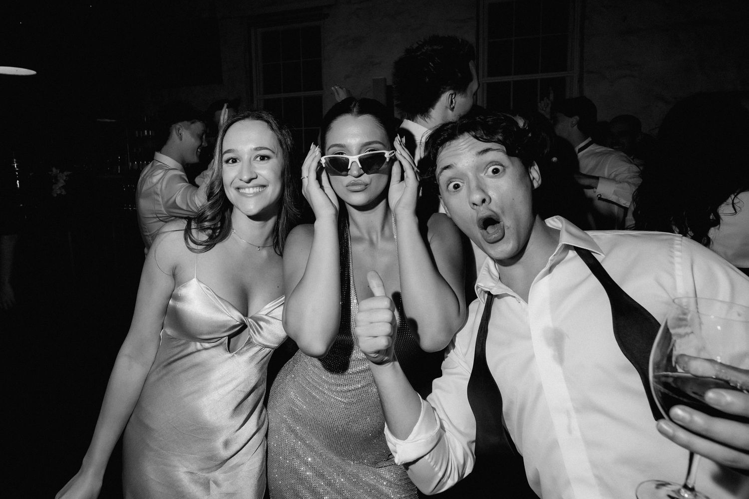 Three friends posing playfully at a wedding reception in black and white, woman in sunglasses giving thumbs up.