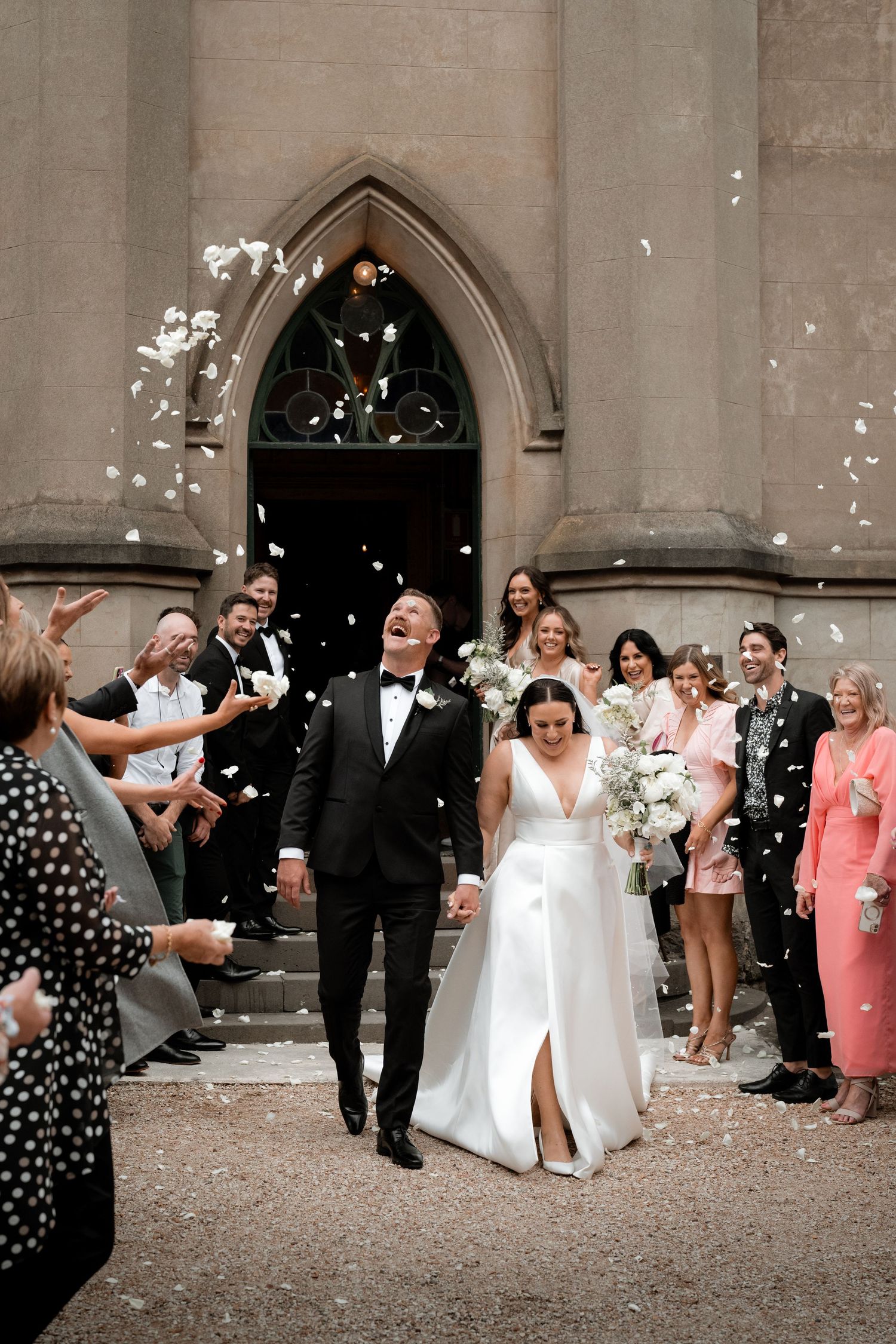 Newlyweds walk down church steps while guests celebrate by throwing white confetti petals outside Gothic archway.