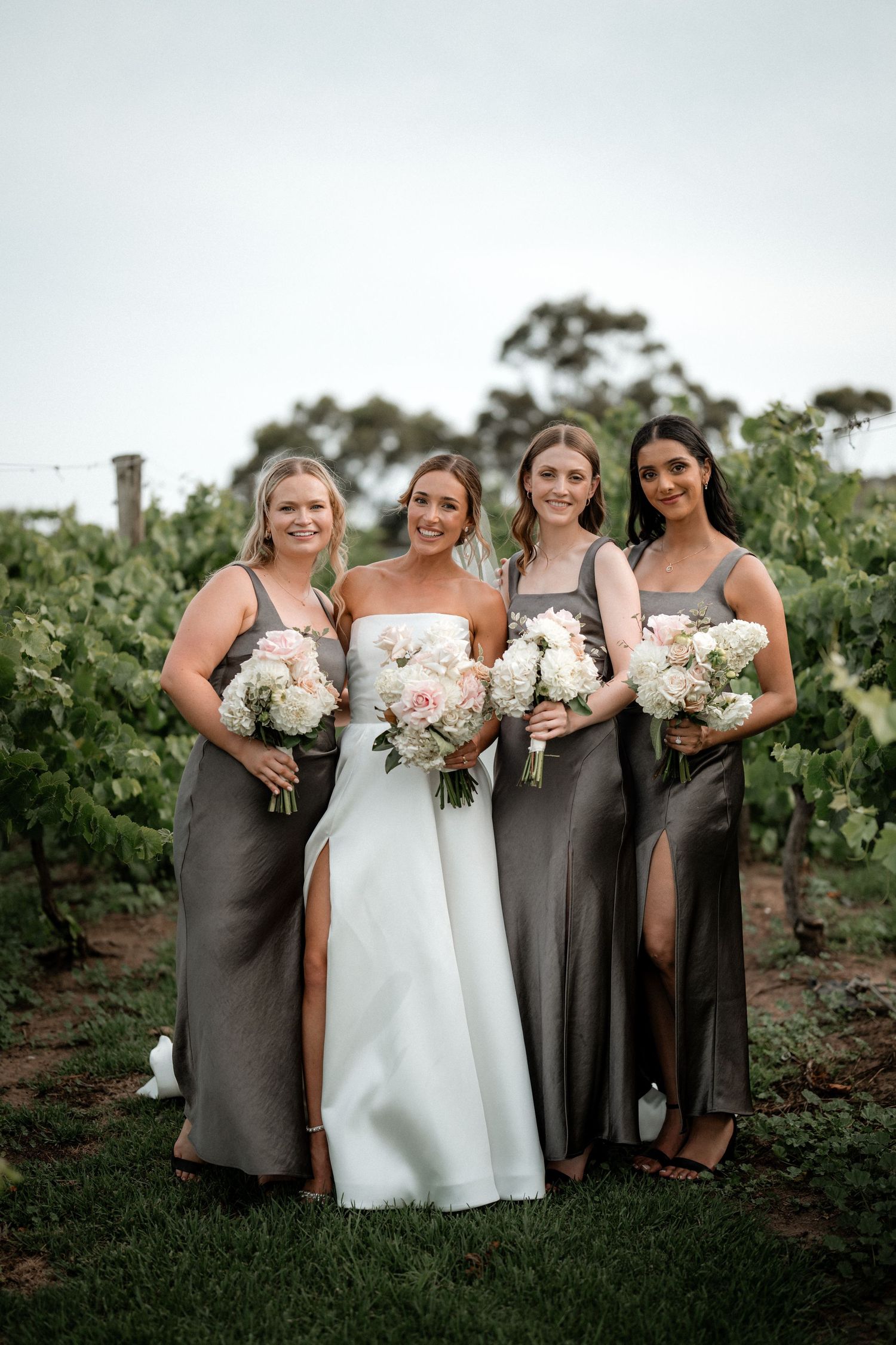 Bridal party in elegant gray dresses and white gown pose with floral bouquets at a vineyard during sunset.
