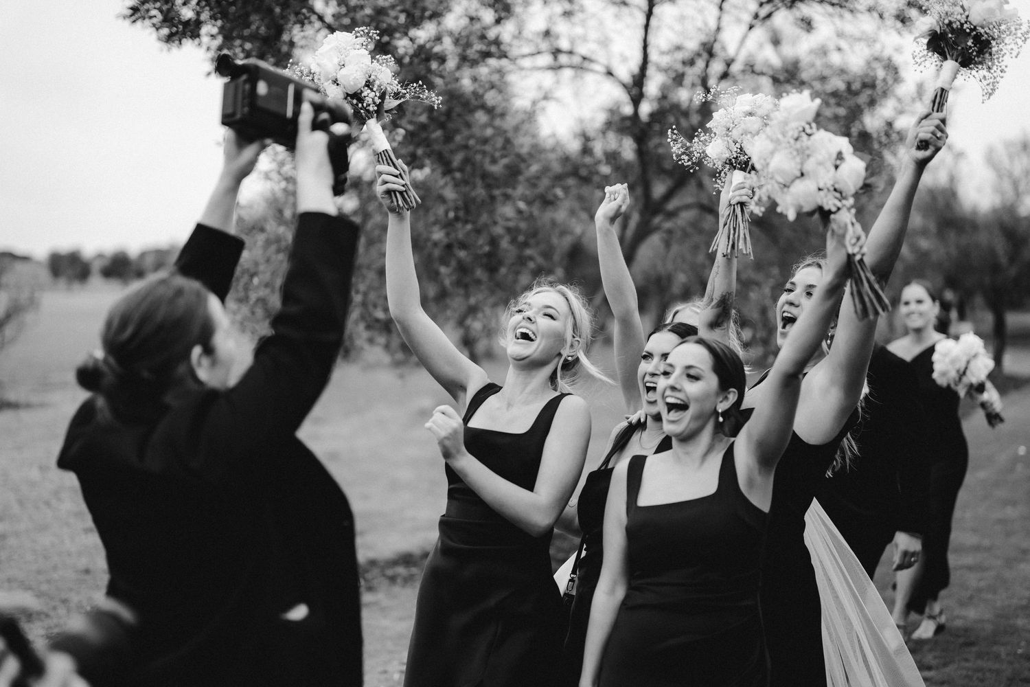 Wedding party members in black dresses celebrating joyfully with raised arms outdoors.