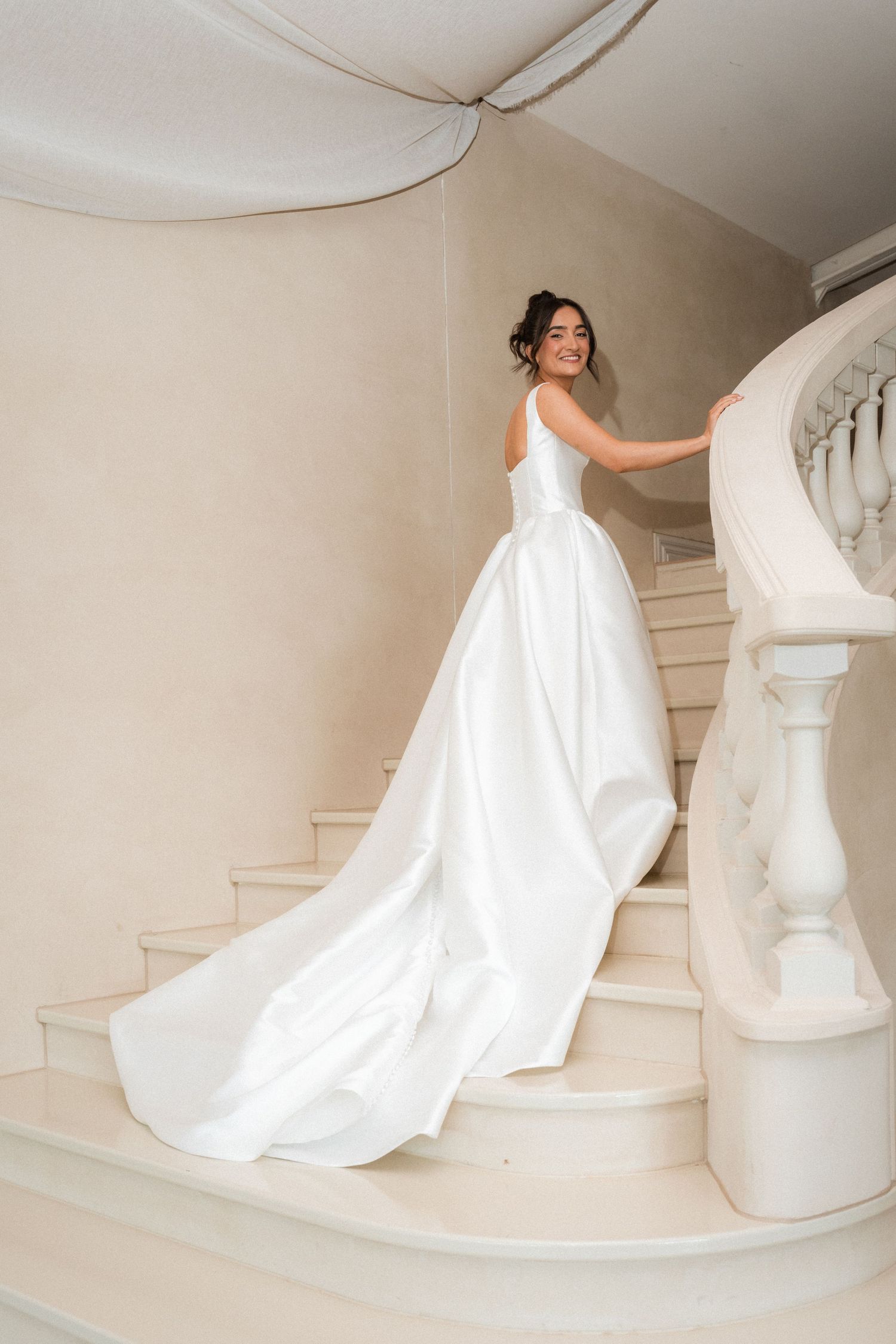 Bride in an elegant white satin ball gown with a long train smiles while ascending a curved marble staircase.