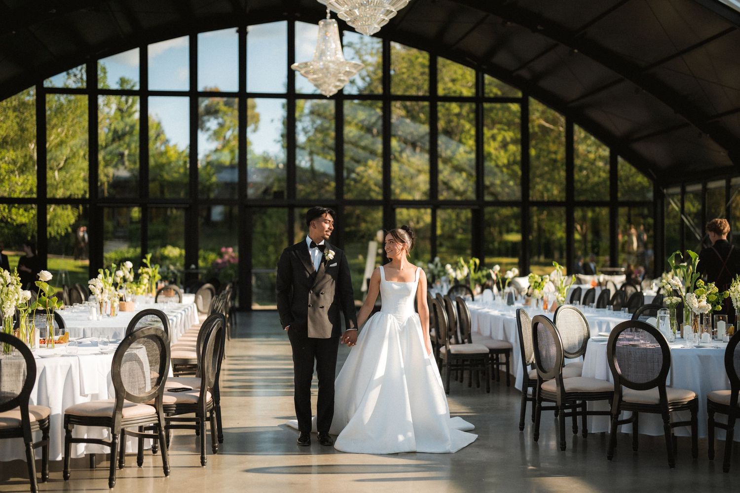 Bride in a white gown and groom in a dark suit walk hand in hand through an elegant glass-walled wedding reception venue with set tables.