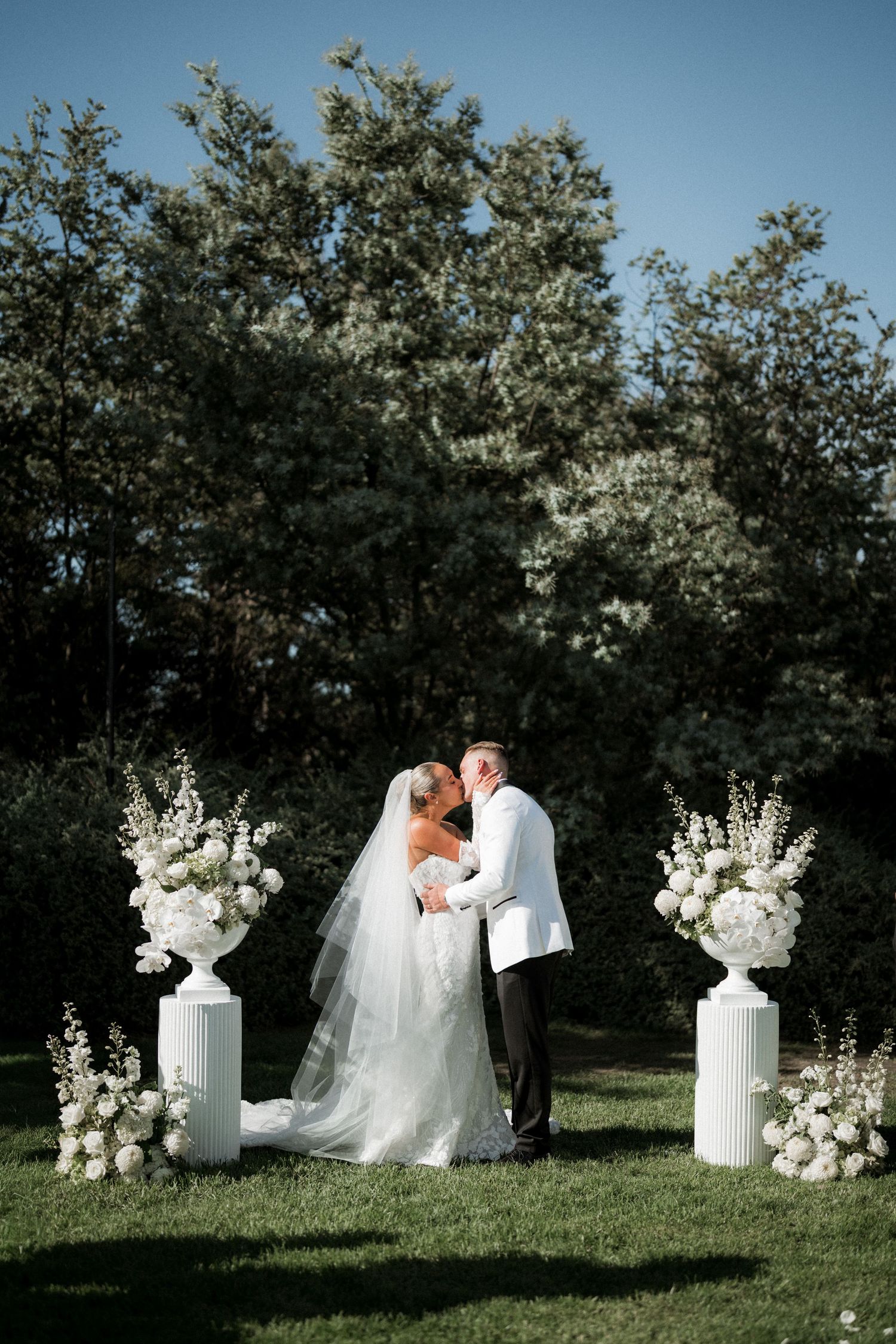 Elegant outdoor wedding ceremony setup with white floral arrangements on pedestals and a couple sharing an intimate moment.