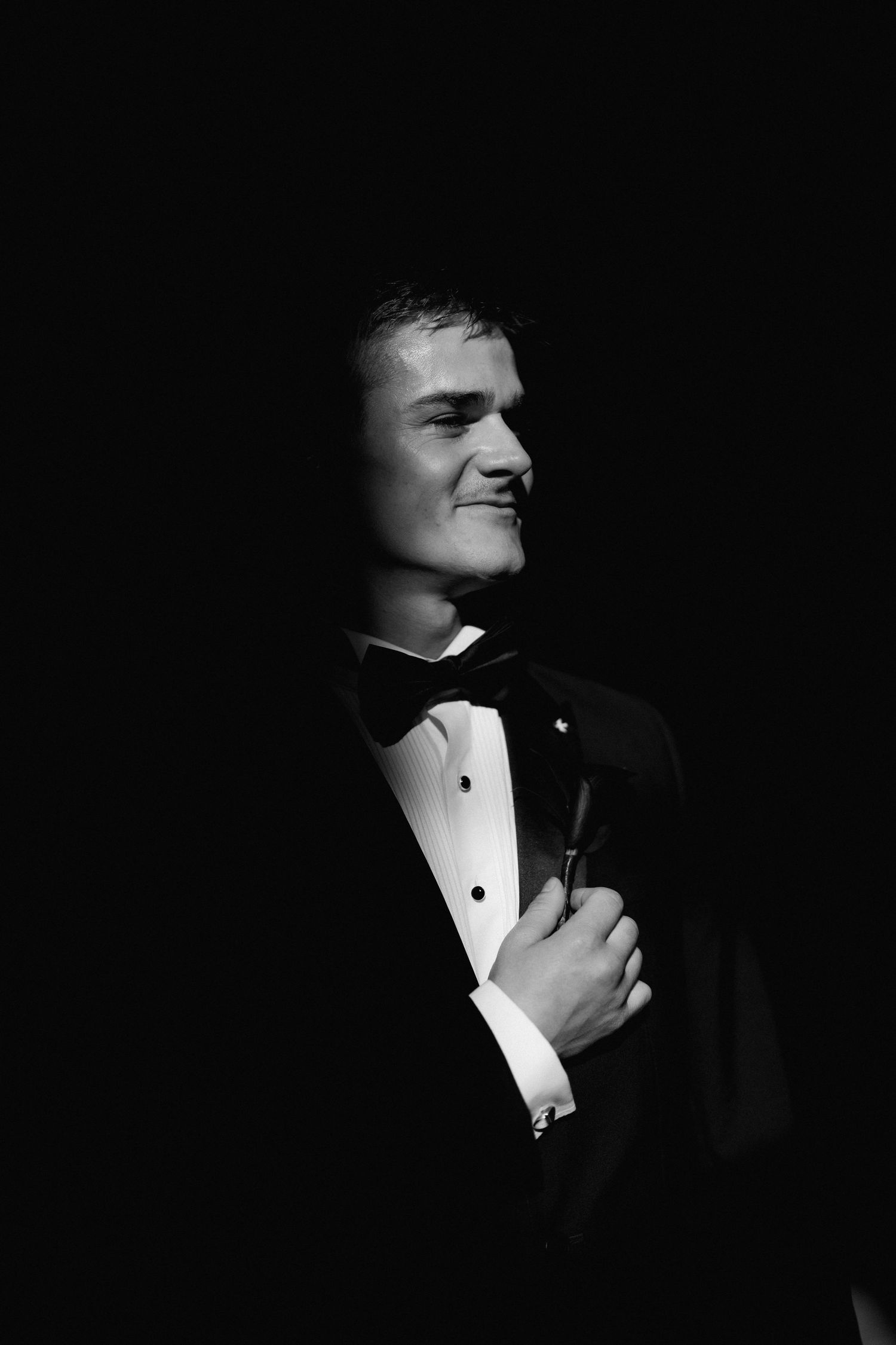 A dramatic black and white portrait in low key lighting shows a person adjusting their formal bowtie.