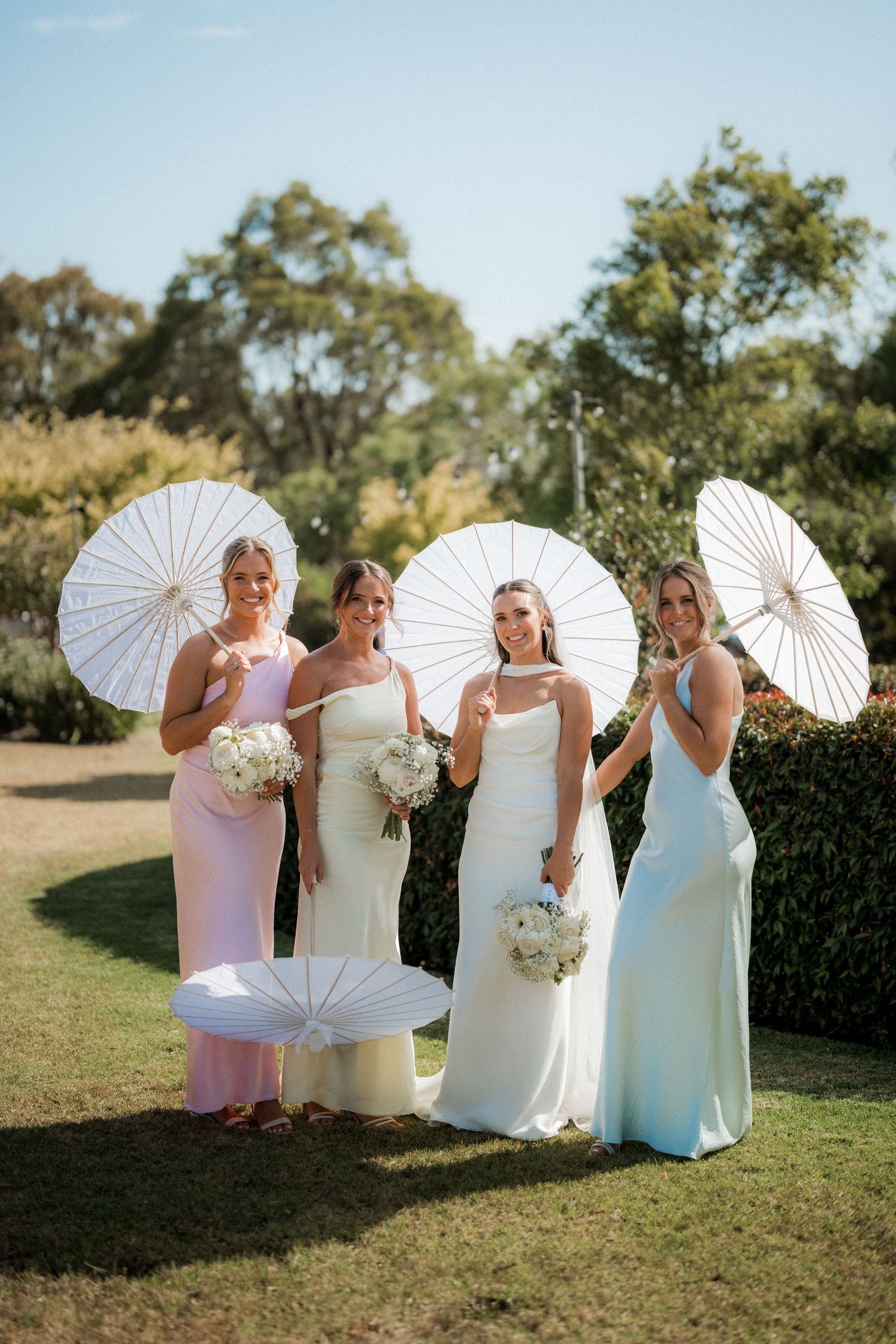 Bridesmaids in pastel dresses pose with white parasols on a sunny lawn during an outdoor wedding.