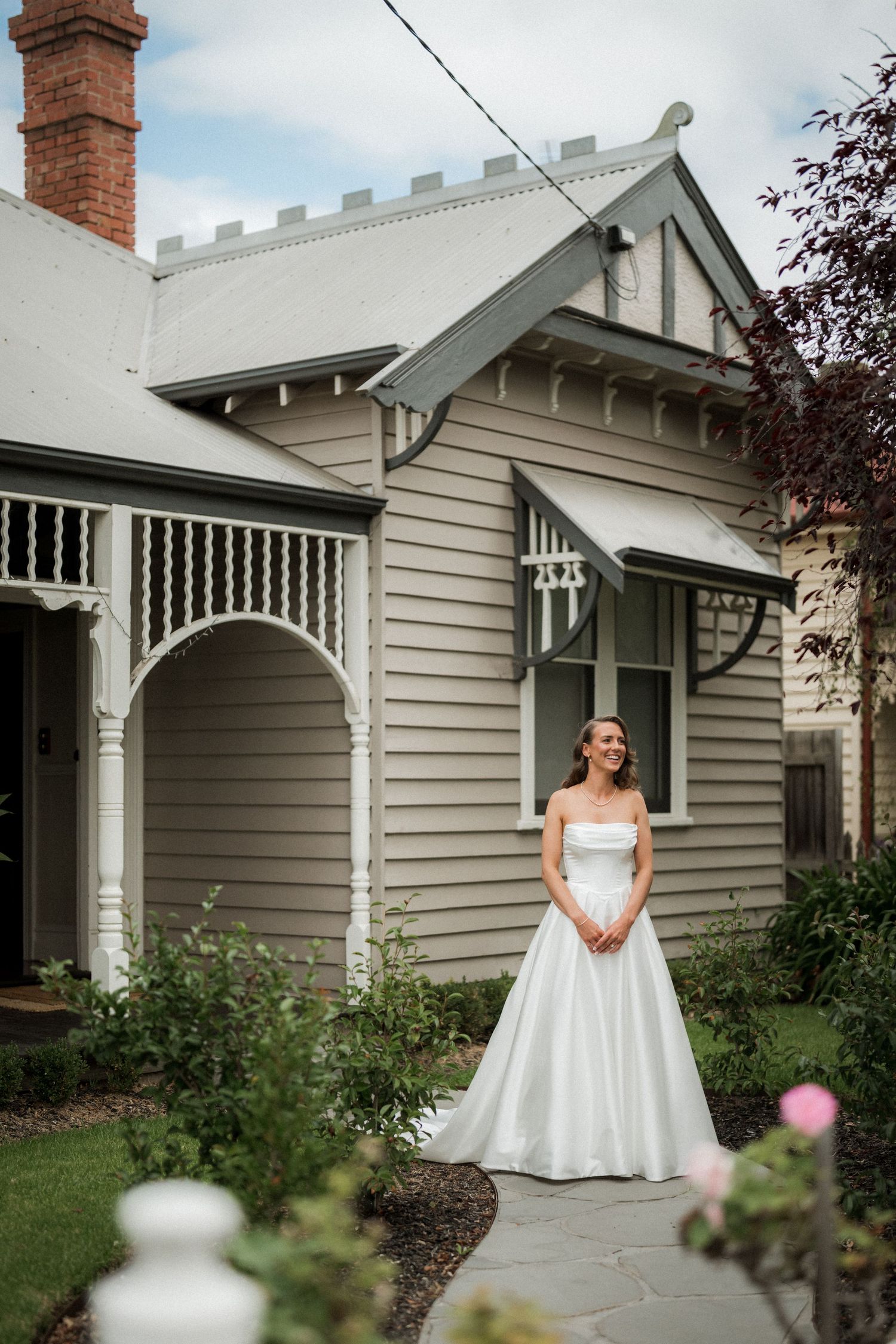 Bride in a classic white wedding dress poses in front of a charming weatherboard house with brick chimney.