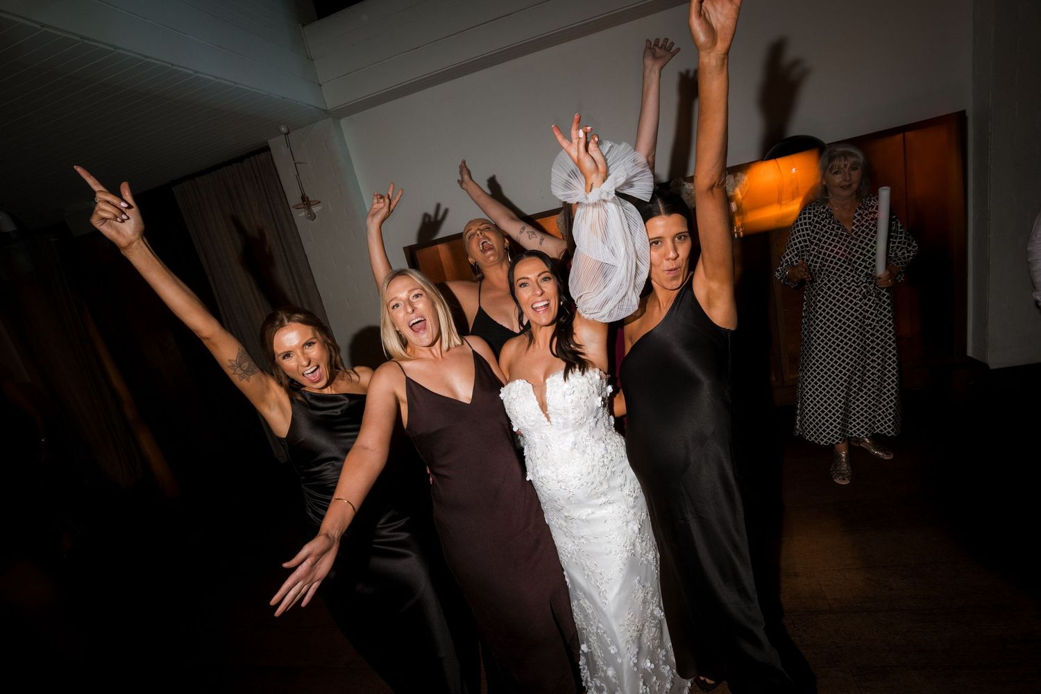 Wedding party dances and celebrates with raised arms in a dimly lit reception venue.