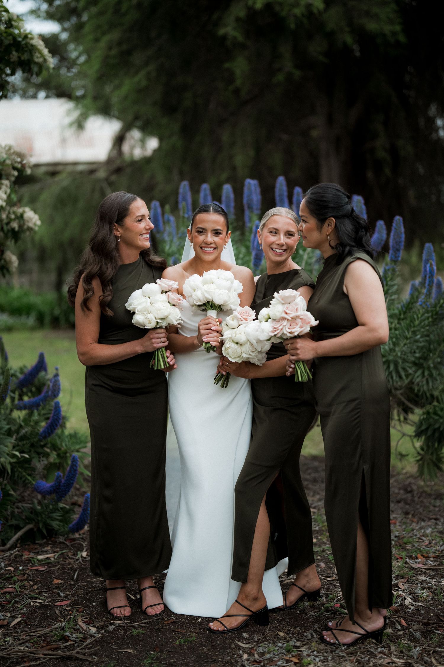 Wedding party poses with white bouquets against garden backdrop with blue delphiniums.