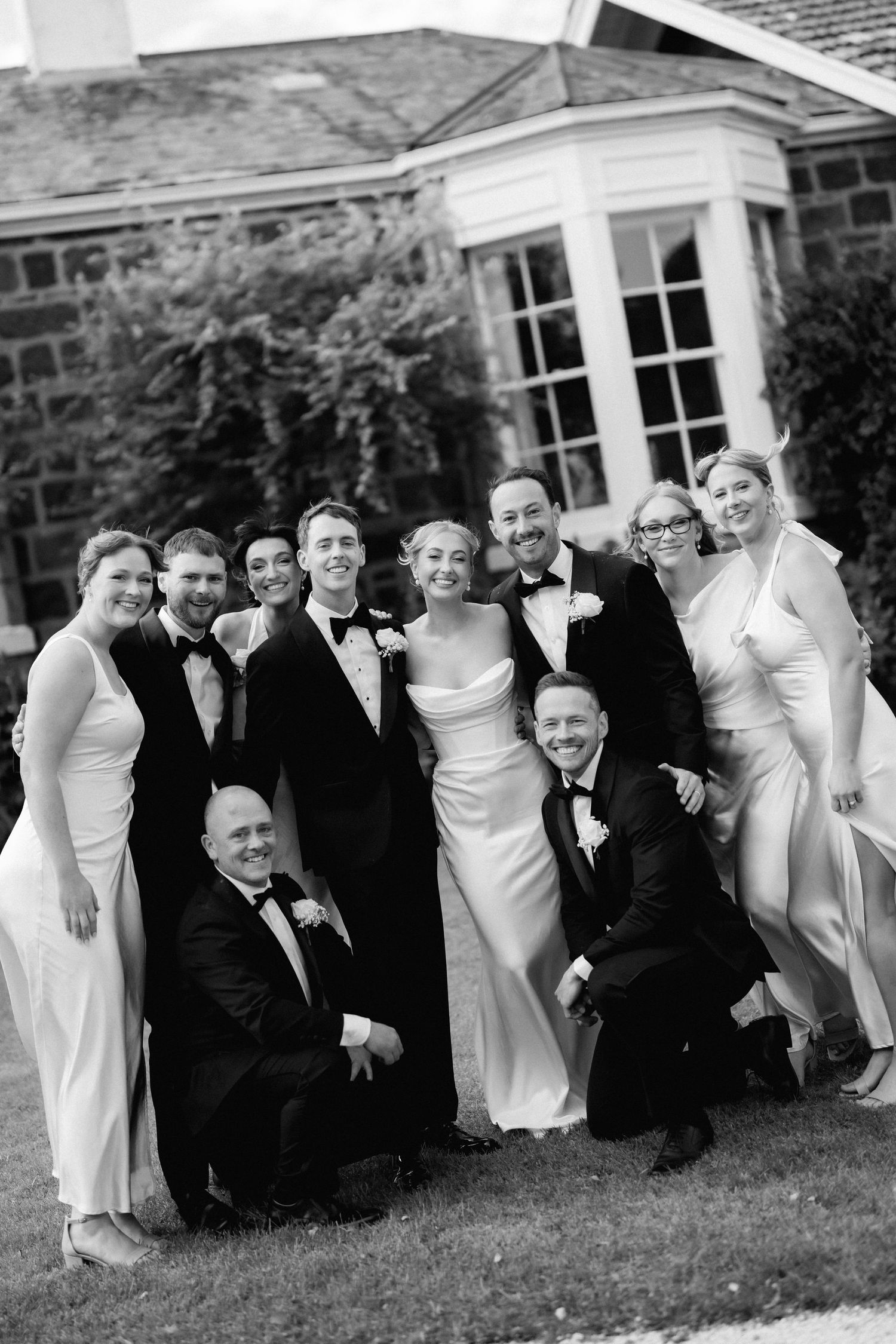 Wedding party in black and white attire posing together on the lawn of a stone building.