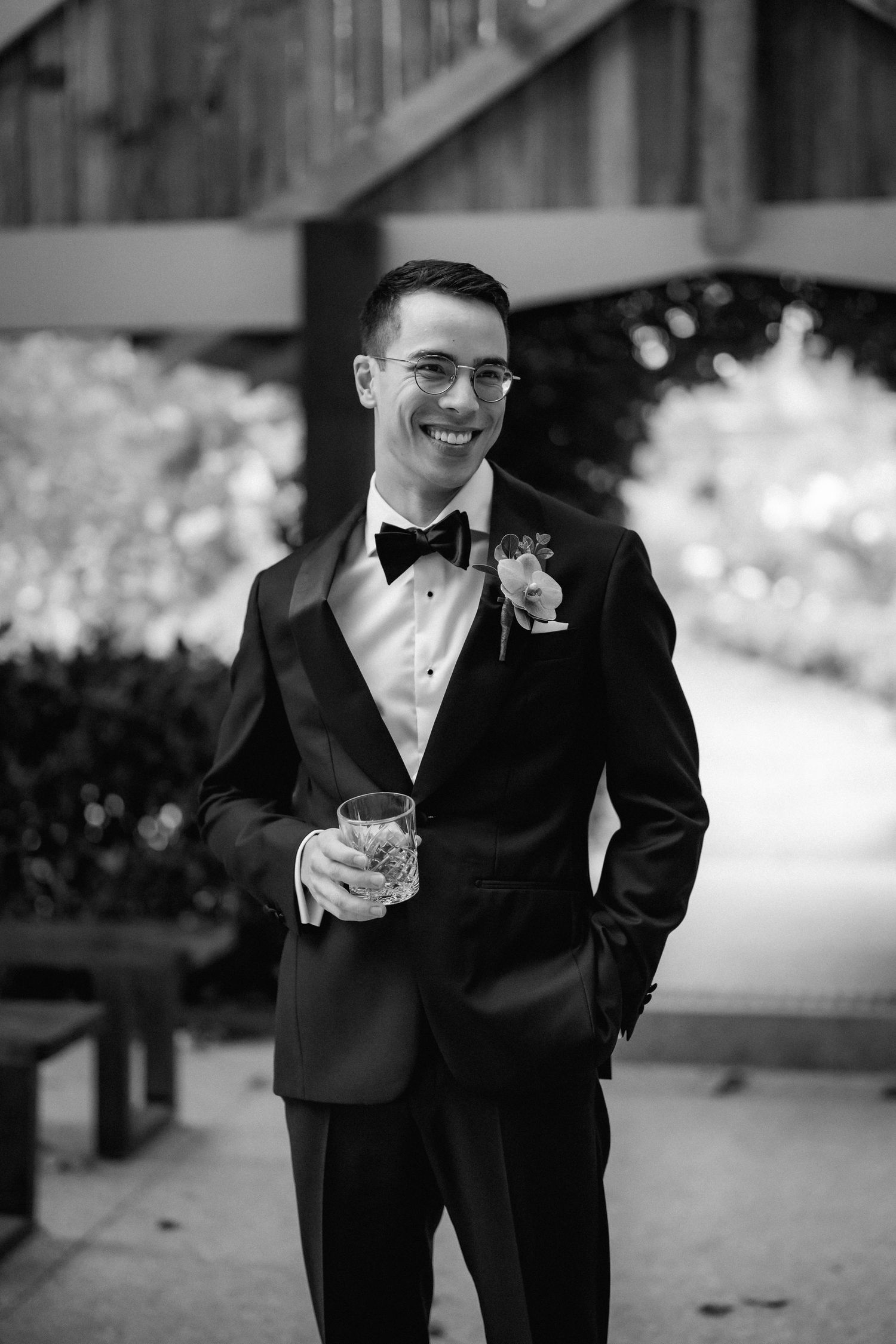 Smiling groom in black tuxedo with bow tie holding whiskey glass at outdoor wedding venue.